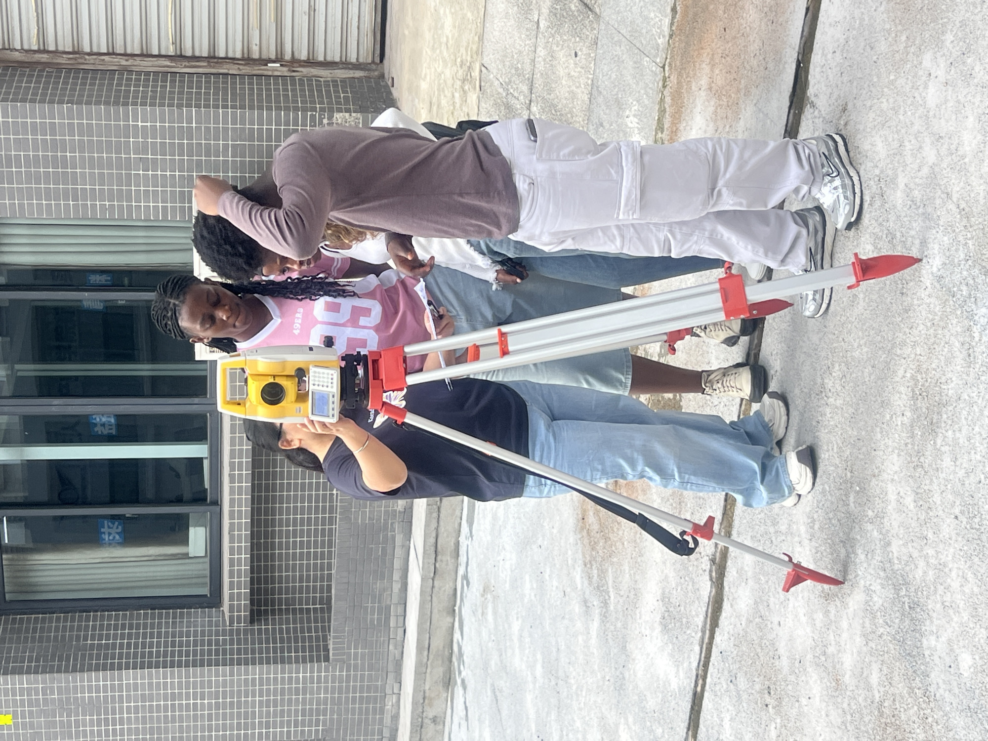 The image depicts four individuals on an exterior concrete surface in what appears to be a city environment. A gray and white tiled building with multiple windows is visible in the background. The focal point is a yellow surveying instrument mounted on a red and silver tripod, with one individual, wearing a black t-shirt and light blue jeans, seemingly operating it. A person in a pink jersey with the number "86" is standing near the tripod. Two additional individuals, one in a lavender long-sleeved shirt and white cargo pants and the other in a white shirt and blue jeans, are standing to the side and looking down. The location is Jiangjin, China.
