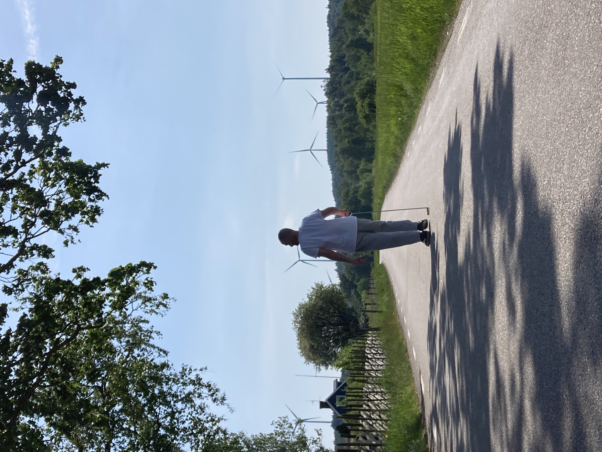 A bald man, wearing a white t-shirt and grey trousers, stands on the right side of a paved asphalt road in a sunny, rural landscape in Vessigebro, Sweden. He is viewed from a slightly rear angle, with his head turned towards the left, and is holding a stick, possibly a walking stick or a golf club, vertically with his right hand.
The road extends from the lower right of the frame towards the upper left, featuring a white line marking its edge. Prominent, jagged shadows from trees fall across the road, suggesting strong sunlight. To the immediate left of the road is a green grassy verge, followed by a rustic wooden fence. Beyond the fence, and also to the right side of the road, are dense green trees and foliage, indicative of a forested environment.
In the mid-ground and distant background, several large wind turbines are visible, their blades stark against a clear, bright blue sky with only a few wispy clouds. These turbines are interspersed among rolling, forested hills. The scene suggests a pleasant day with bright weather, likely midday or early afternoon given the distinct and relatively short shadows. No text is visible in the image. The overall setting combines natural beauty with elements of renewable energy infrastructure.