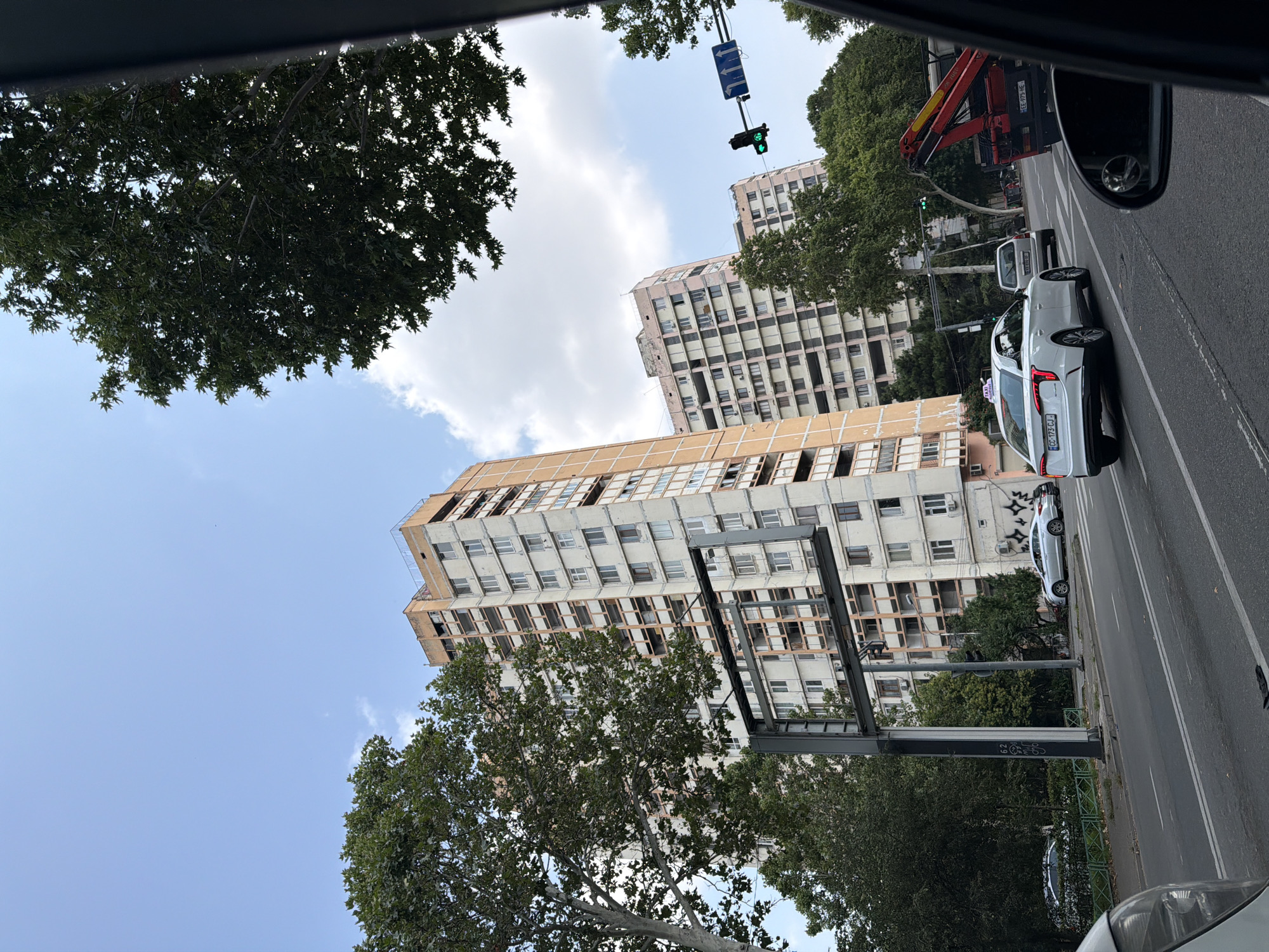 The image captures a street scene in Tbilisi, Georgia, featuring several high-rise apartment buildings. The buildings appear to be of Soviet-era architecture, characterized by their concrete facades and multiple balconies. The sky is partly cloudy, with a mix of blue sky and white clouds. Trees with lush green foliage are interspersed between the buildings and along the street, suggesting a relatively warm season.
In the foreground, a white taxi is visible on the road, indicating urban activity. Other vehicles, including a red tow truck, are also present, suggesting traffic flow. A traffic light is illuminated green, further emphasizing the ongoing movement of vehicles. The road itself is paved and appears to be a multi-lane street. The overall impression is of a typical urban environment in Tbilisi, with a blend of residential architecture, greenery, and vehicular traffic. The image was taken from inside another vehicle, as indicated by the partial view of a car's interior elements, such as a side mirror.
