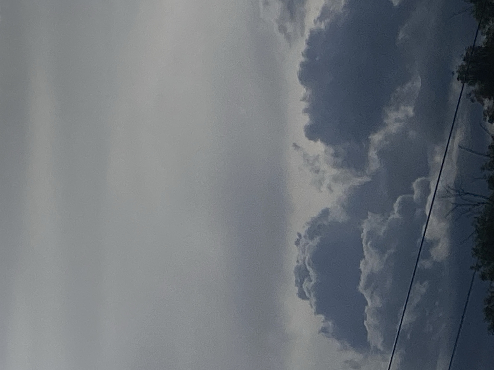 The image shows a cloudy sky with a mix of overcast and cumulus clouds. A power line runs diagonally across the right side of the frame. Some foliage is visible on the far right, possibly trees. There are no people or discernible objects. The location is possibly Заволжье, Russia.
