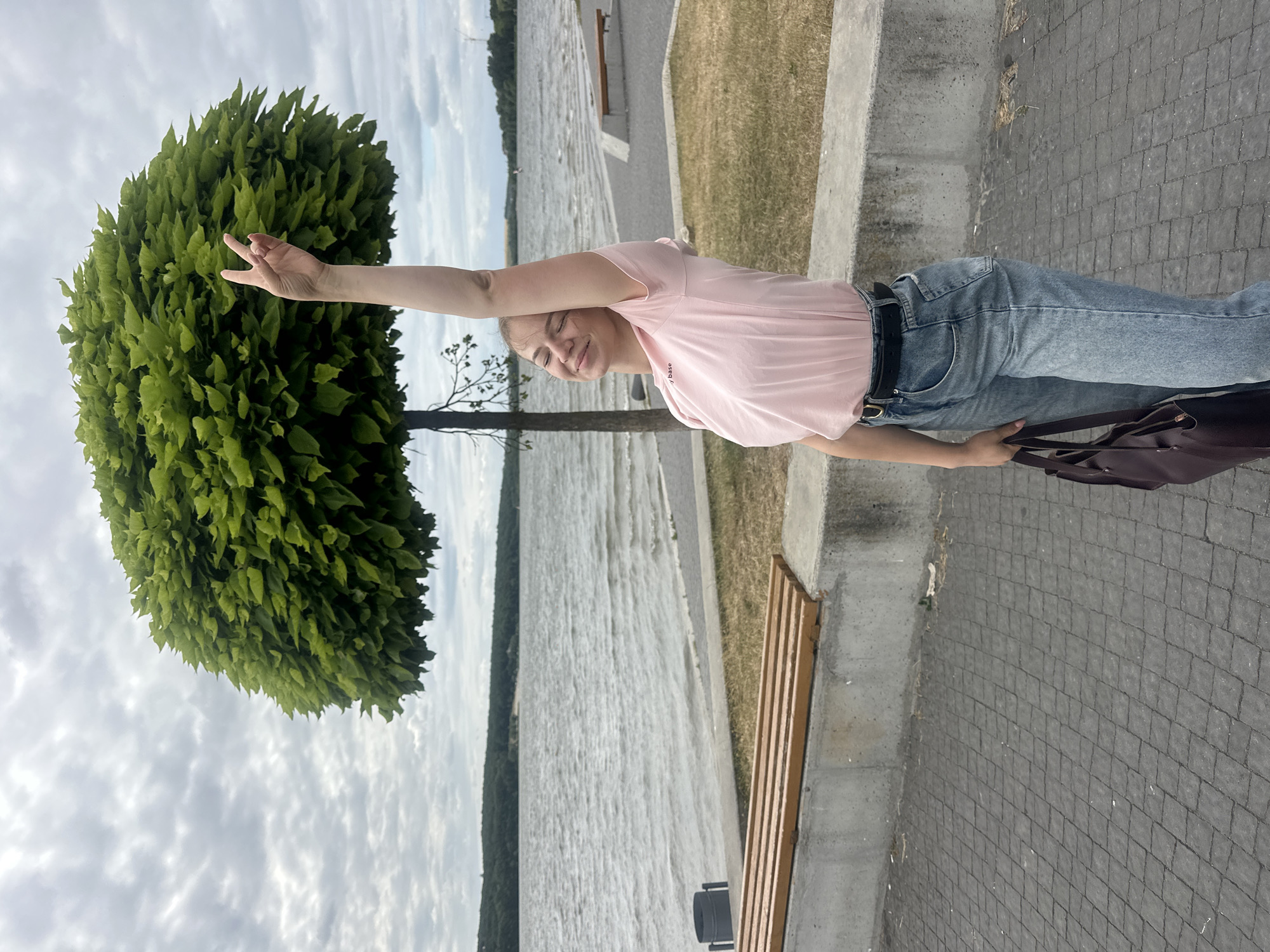 A young woman stands on a cobblestone path next to a lake, raising her right arm in a peace sign gesture. She is wearing a pink t-shirt, blue jeans, and a black belt. She is holding a dark brown bag in her left hand. The woman has her eyes closed and a slight smile on her face. Behind her, a body of water stretches towards a distant treeline under a cloudy sky. A trimmed, round green tree is positioned to her left. In the foreground, there are two wooden benches. The scene appears to be a public waterfront area in Ternopil, Ukraine.