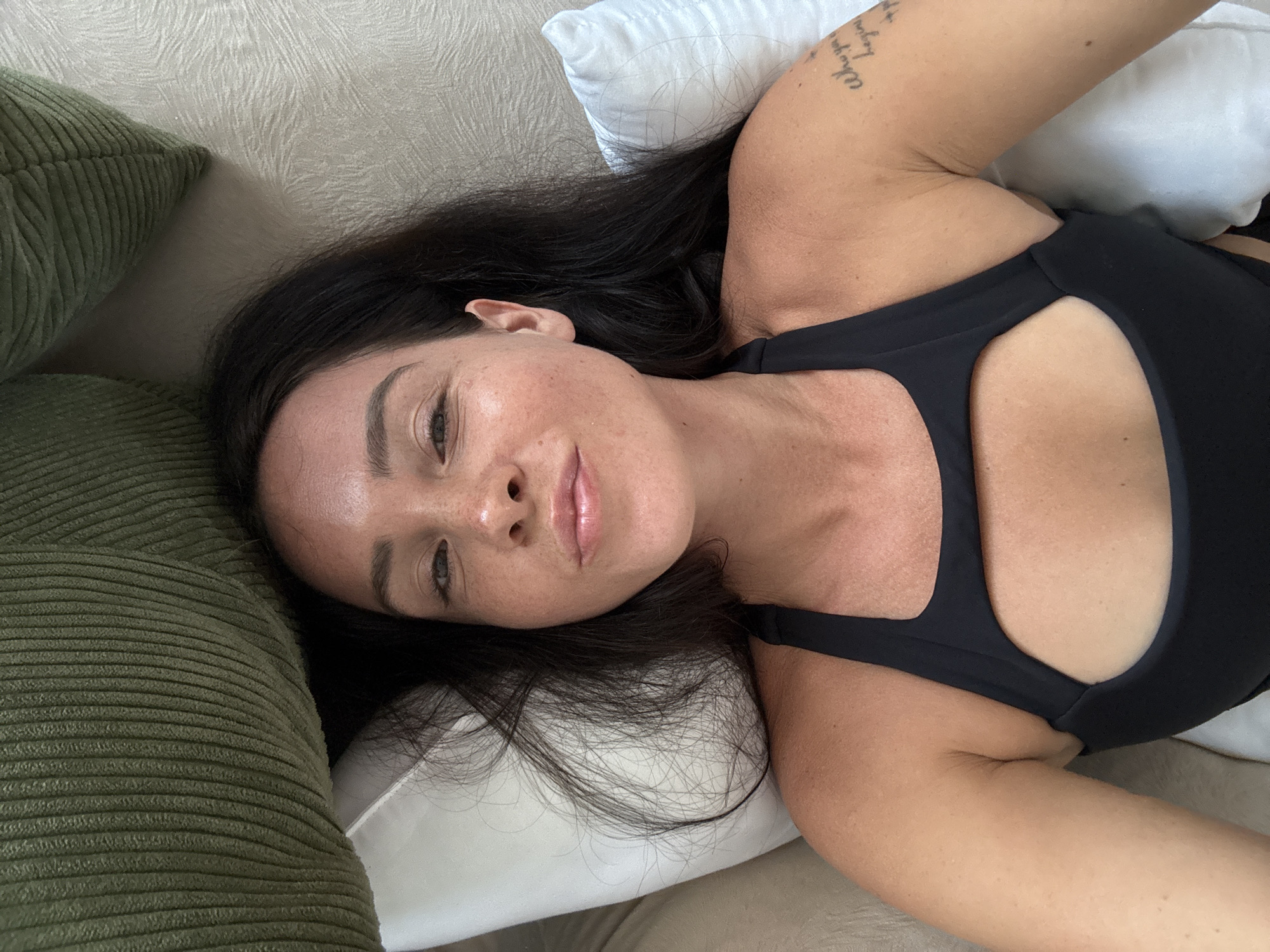 A young woman with long, dark hair is lying on a bed, looking up at the camera. She is wearing a black sports bra with a cutout in the chest area. Her skin has a slight tan and is dotted with freckles. Her lips are slightly parted and appear to be glossy. She has a tattoo on her left arm. The bed appears to be made of white linen, and there are pillows and a green corduroy pillow in the background. The setting is likely a bedroom in North Kuta, Indonesia, given the lighting and overall ambiance.