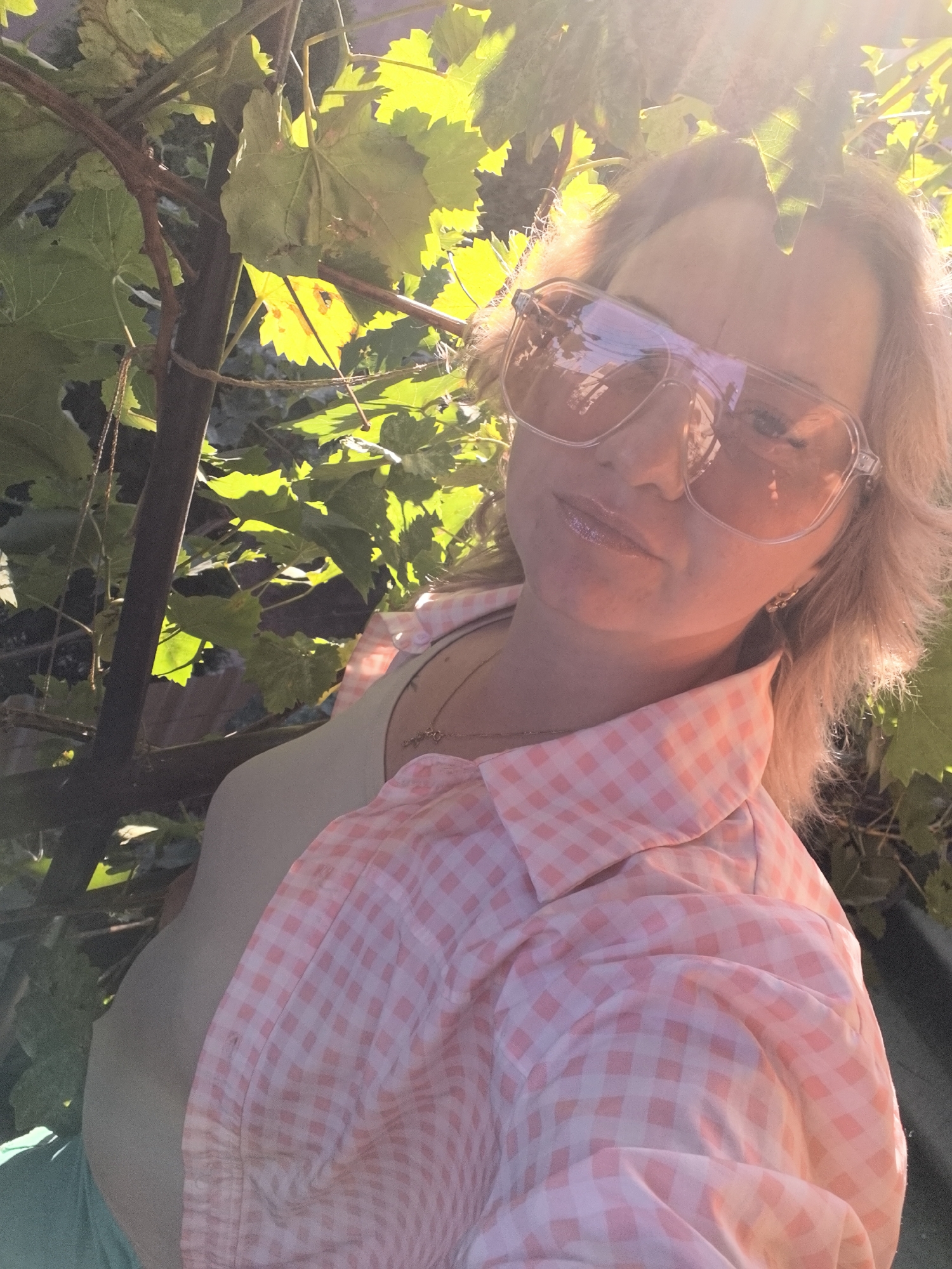 A woman is pictured outdoors, close-up, wearing large, clear-rimmed sunglasses with a pink tint and a pink and white gingham collared shirt. She has blonde hair and appears to be smiling with glossy lips. The background consists of green leaves and branches of a vine, illuminated by bright sunlight, suggesting a sunny day. The lighting creates lens flare across the top of the image. The location is not specified as Vityazevo, Russia, as this information cannot be determined from the image itself.