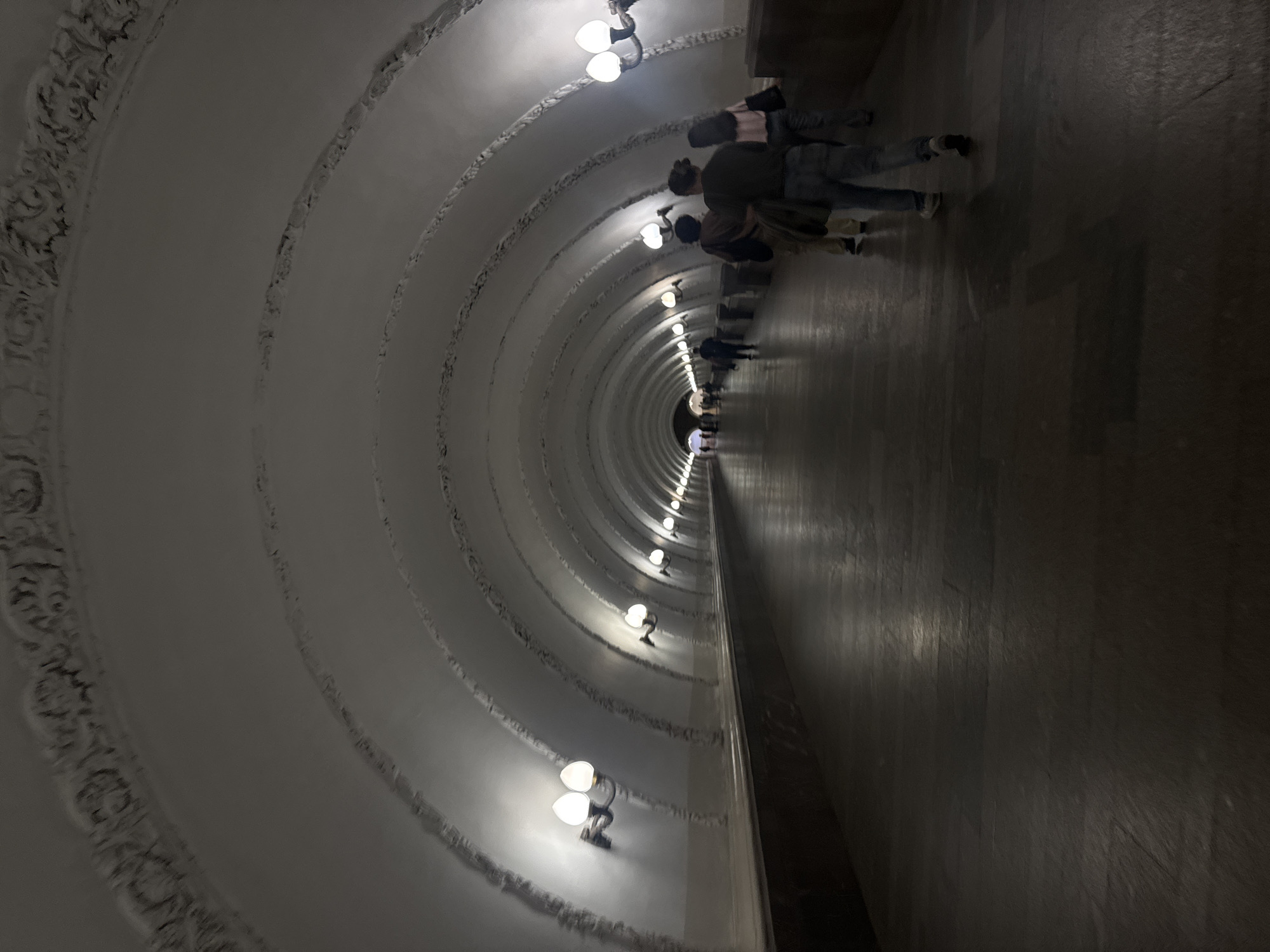 The image shows a long, arched corridor in the Moscow Metro, Russia. The architecture is characterized by repeating arched structures, each adorned with decorative moldings and lit by evenly spaced, old-fashioned lamps. Several people are walking through the corridor, their figures diminishing in size as they recede into the distance, conveying depth. The floor has a dark, polished appearance.