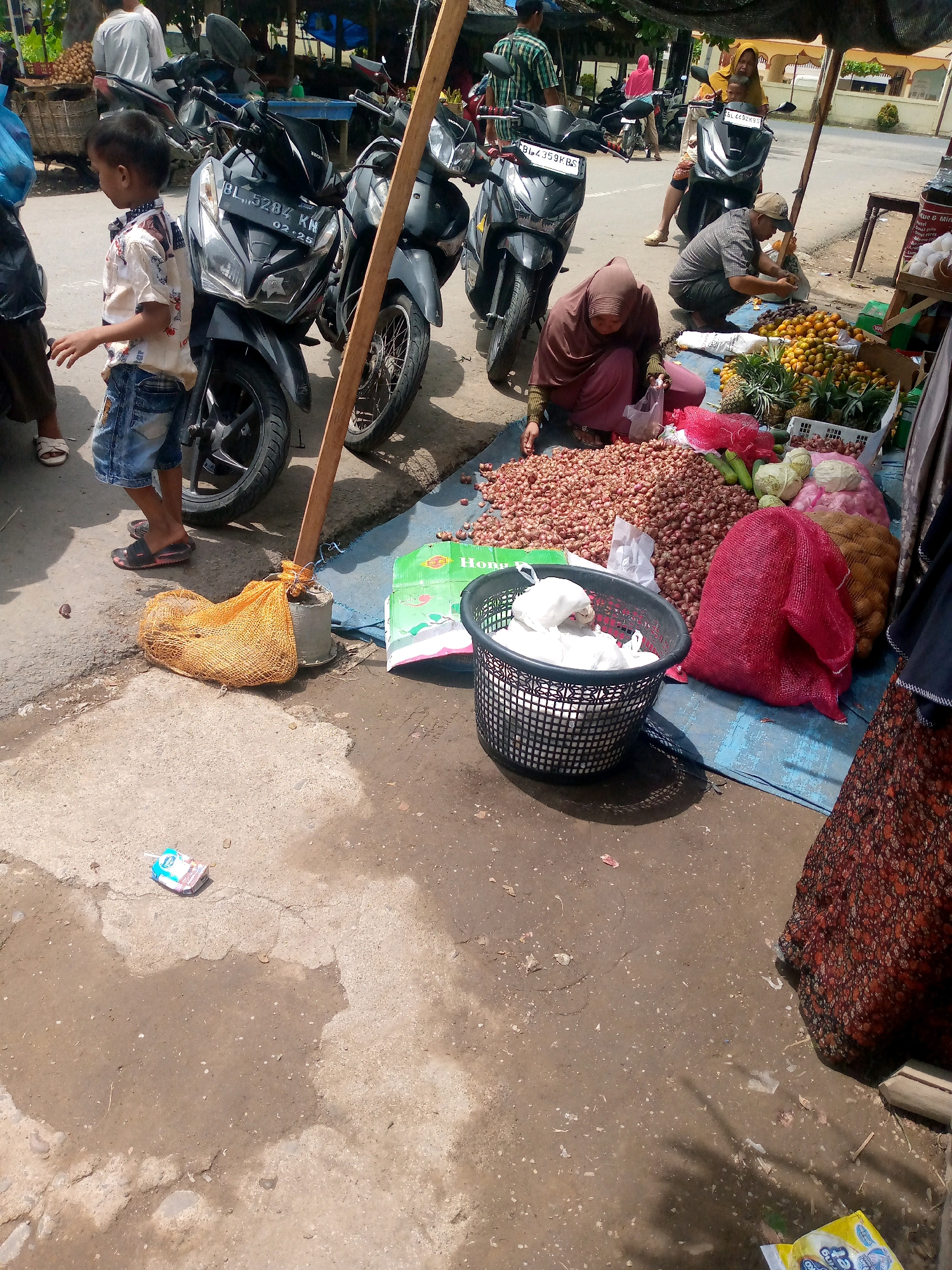 In Keudeteupinpunti, Indonesia, a street scene unfolds with a focus on commerce. A young boy, dressed in a patterned shirt and denim shorts, stands beside a motorcycle. To his right, a woman in a brown headscarf crouches, sorting through a large pile of red onions. In front of her, a black plastic basket contains white items, possibly fabric. Behind the onions, a collection of fruits and vegetables is displayed, including pineapples, oranges, and cabbages, some of which are bagged. Another woman, also in a headscarf, sits on a motorcycle in the background, holding a child. The ground is a mixture of dirt and patches of concrete, littered with small debris. A wooden pole stands prominently in the foreground, partially obscuring the scene. A discarded juice box lies on the ground near the bottom of the image.