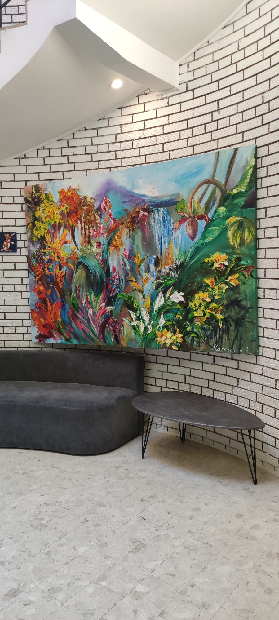 The image depicts an indoor scene featuring a large, vibrant painting of a tropical landscape with a waterfall. The painting is mounted on a white brick wall with dark grout. In front of the painting, a modern, dark gray curved sofa is placed. To the right of the sofa, a dark gray, teardrop-shaped coffee table with thin black legs is situated. The floor is covered in light-colored terrazzo tiles. The overall setting appears to be a contemporary indoor space, possibly a lounge or waiting area. There are no people visible in the image. The location is stated to be in Rosinka, Russia.