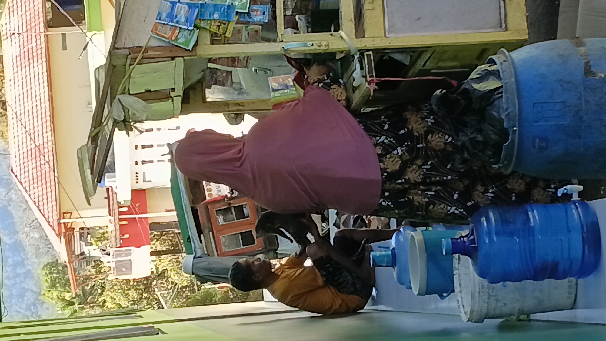 In a scene set in Mandalaguna, Indonesia, a man wearing an orange t-shirt and dark shorts sits on a ledge, facing away from the viewer. He is holding a dark object, possibly a cat, in his lap. To his right, a woman in a pink hijab and a patterned skirt stands behind a makeshift stall. The stall appears to be constructed from various materials, including wood and corrugated metal, and displays an assortment of items, including packaged goods and what seem to be water containers. A prominent blue water jug is visible in the foreground, along with other buckets and barrels. In the background, a building with a red-tiled roof and white walls is visible, surrounded by lush greenery and a mountainous landscape. The overall impression is of a casual outdoor commercial space, possibly a roadside stall or a small shop.