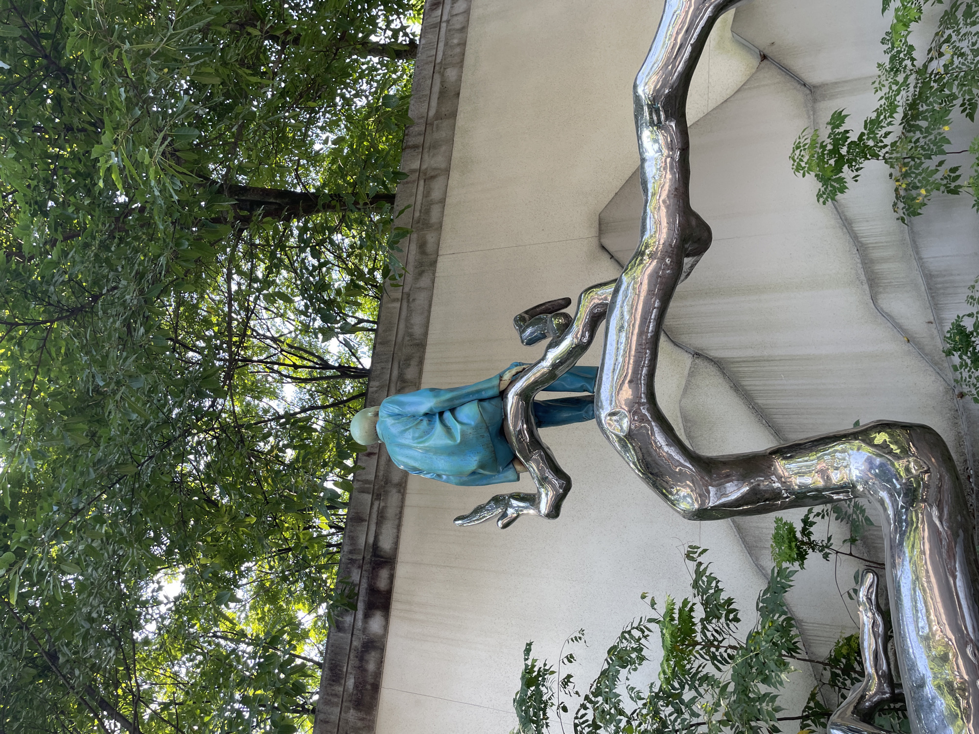 The image shows a sculpture of a person sitting on a branch. The sculpture is made of polished metal and has a blue-colored figure sitting on top of it. The figure is facing away from the camera. The branch is a part of a larger, abstract metal sculpture that curves and twists. The background consists of a textured wall with some greenery in front of it, and trees and foliage above. This sculpture is located in Nan'an District, China.