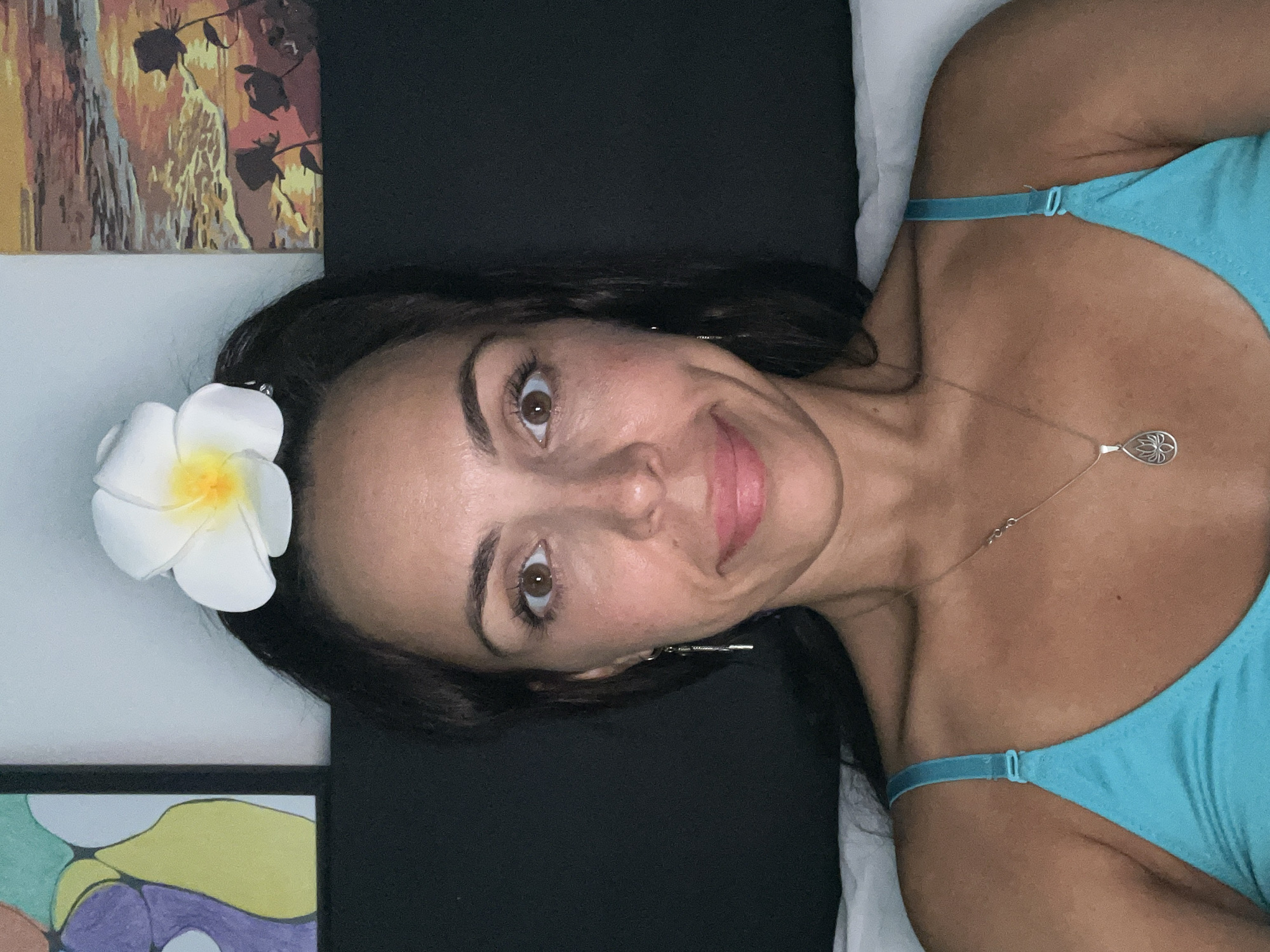 A woman with dark hair is reclining and looking into the camera. She is wearing a light blue tank top and a necklace. A white plumeria flower is placed in her hair. Behind her is a black headboard and a white pillow. Two paintings hang on the wall behind the headboard. The location is inferred to be Kuta Selatan, Indonesia.