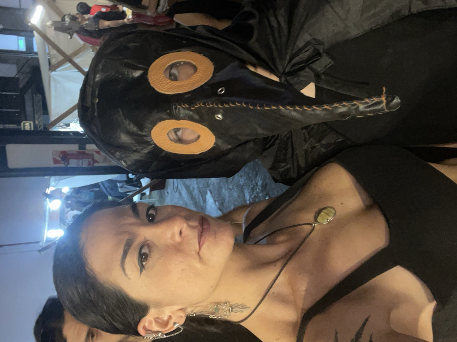 A woman with dark hair and winged eyeliner is visible, wearing a black dress and a pendant necklace. She has multiple earrings on her left ear. Behind her is another person wearing a black plague doctor mask with orange eye holes and a long beak. The scene appears to be at an outdoor event, with tents and other people in the background. The photo is taken at a slightly angled perspective.