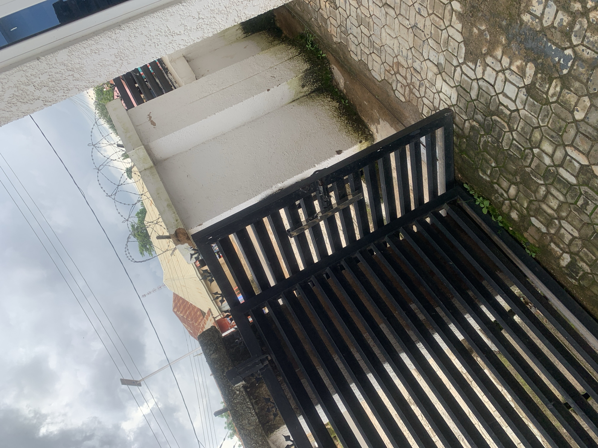 An outdoor, medium shot shows a black metal gate in front of a white, textured wall. The gate is made of horizontal metal bars and is partially closed. The wall has some green moss and dirt at its base. Above the wall, barbed wire is visible, along with several buildings with red and beige roofs, and power lines against a cloudy sky. The ground in front of the wall is paved with interlocking stones that appear wet and dirty.