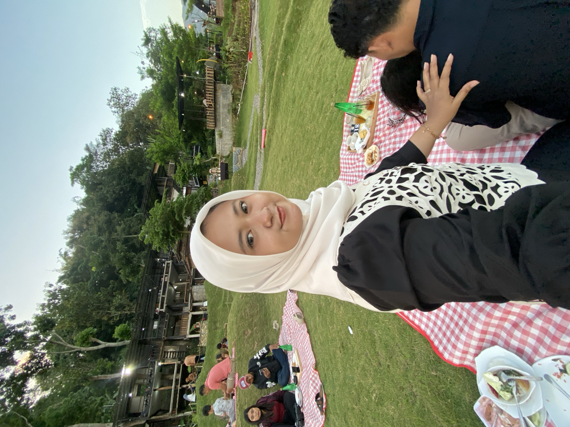 An overhead shot shows a picnic blanket with red and white checks spread on a grassy hill. On the blanket, a small wooden tray holds a bottle of green liquid, two glasses of amber liquid, and a plate of pastries. A woman in a white hijab and black and white patterned top faces the camera and smiles, her left hand resting on the shoulder of another person, whose back is to the camera. Behind them, a man in a dark blue shirt sits facing away. In the background, several other people are seen picnicking on blankets, and beyond them, there are multi-story buildings and lush green trees, suggesting a park or recreational area on a hillside.