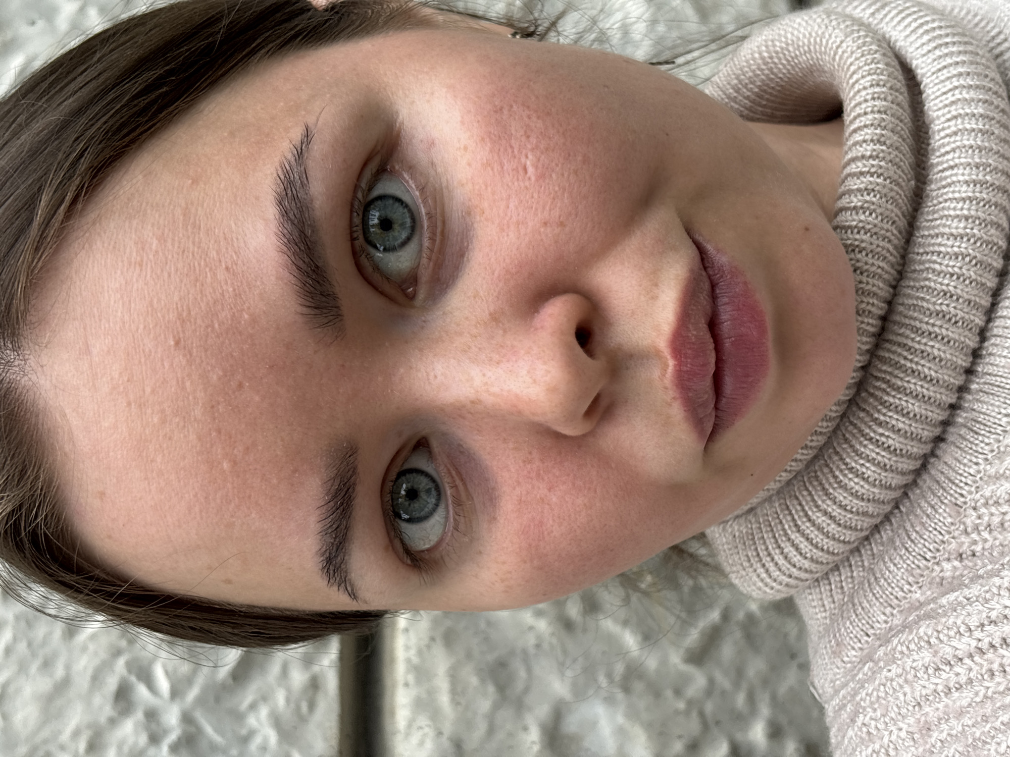 A close-up, slightly angled portrait of a woman with fair skin, blue eyes, and brown hair is centered in the frame. She is wearing a light beige turtleneck sweater. Her expression is neutral, with a slight hint of a smile. The background is out of focus and appears to be a textured white surface. The image does not provide information to place the subject in the city of Русский Вожой, Russia.