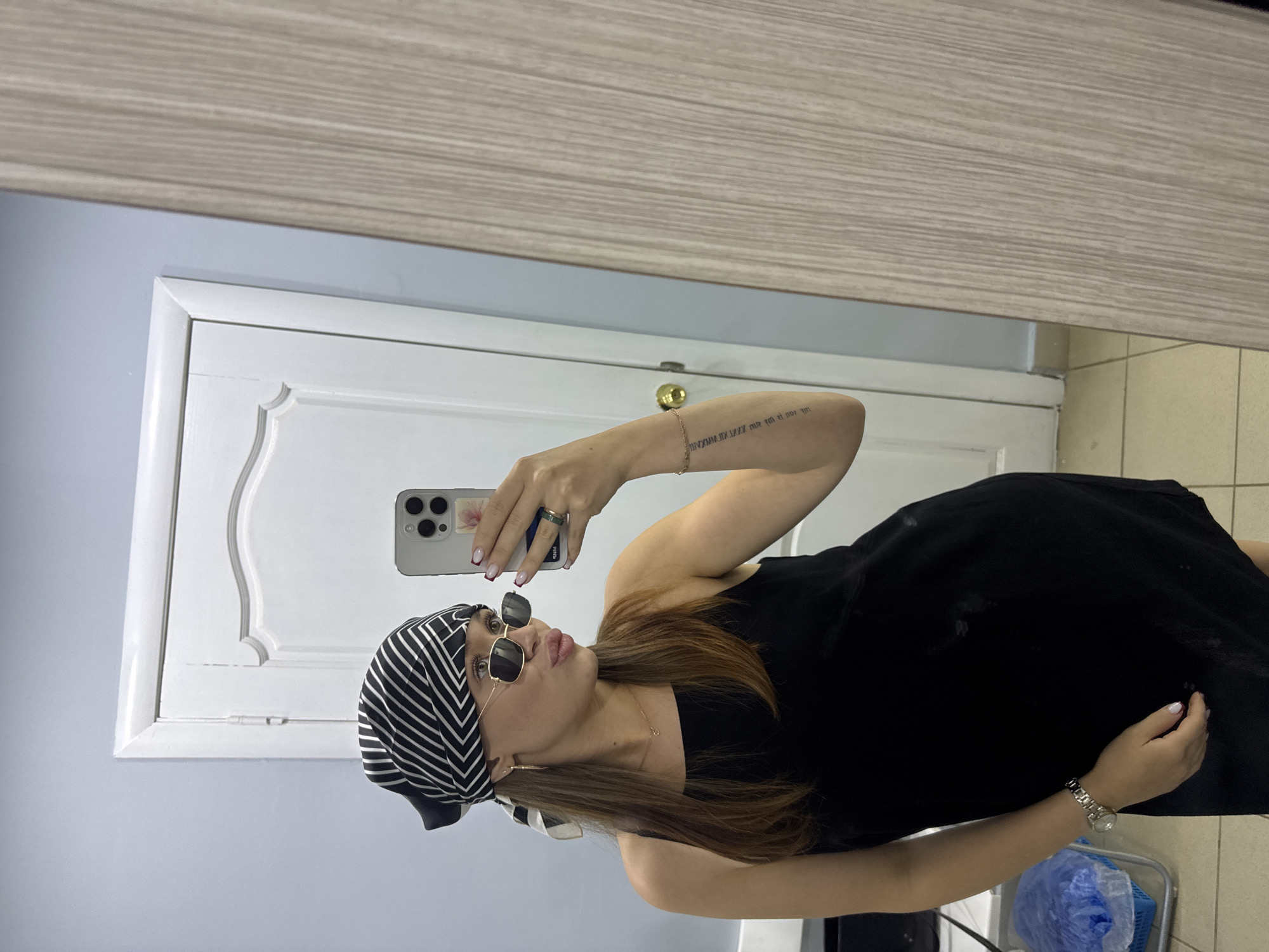 A young woman with brown hair, wearing a black dress and a black and white striped headscarf, takes a mirror selfie. She is also wearing sunglasses, a necklace, a ring, and a watch. Her left arm has a tattoo. The background shows a door and a wall, possibly in an apartment. The presence of a door and the style of the interior suggest a residential setting within the city of Chaikovsky, Russia.