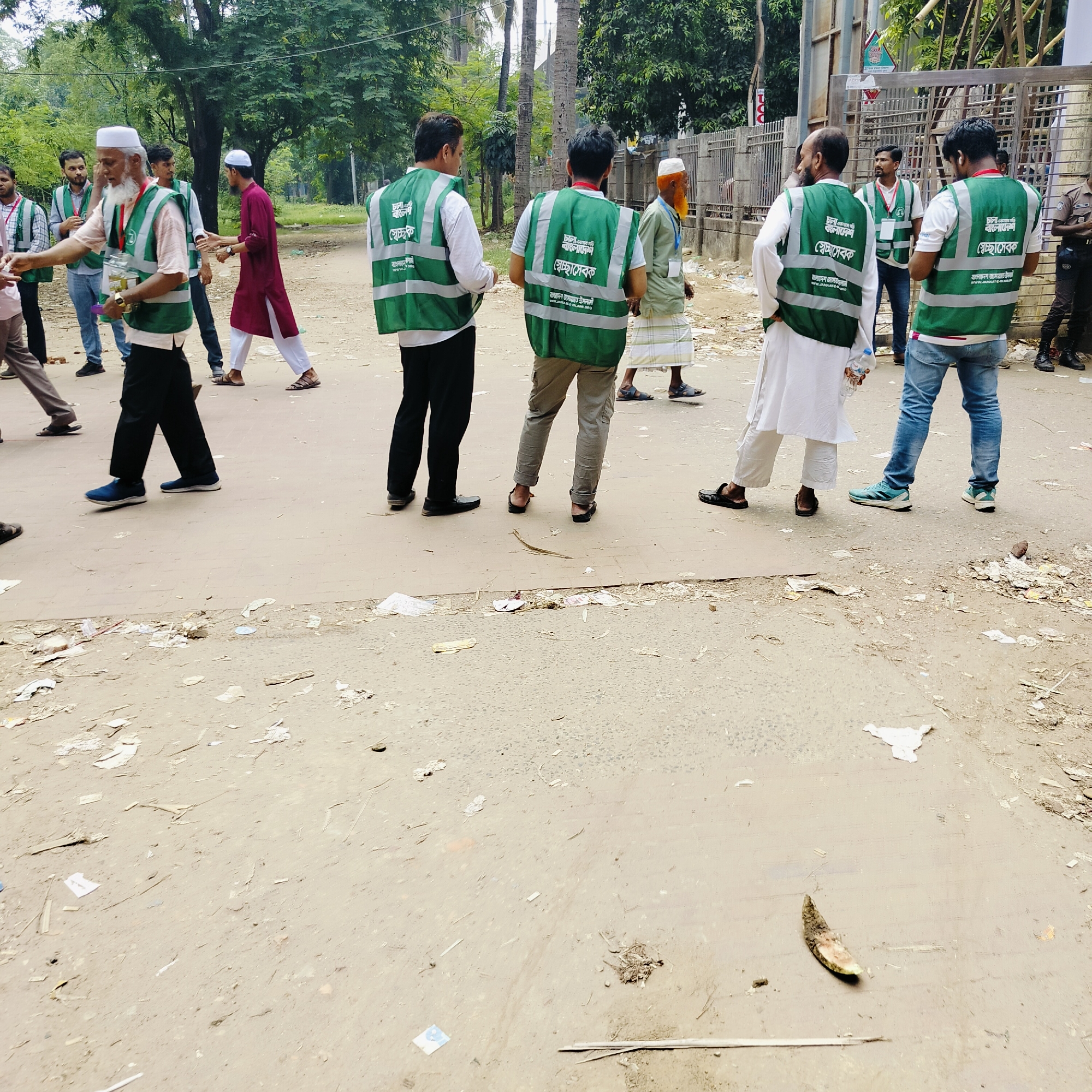 The image shows an outdoor scene in Dhaka, Bangladesh. A group of men wearing green vests are standing and interacting on what appears to be a street or road. The vests have text written in Bengali, along with "স্বেচ্ছাসেবক" which can be translated to "Volunteer." Some men are wearing traditional Muslim attire. The road surface is dirty with scattered litter and debris. In the foreground, there is discarded organic matter. The environment consists of trees and buildings in the background.