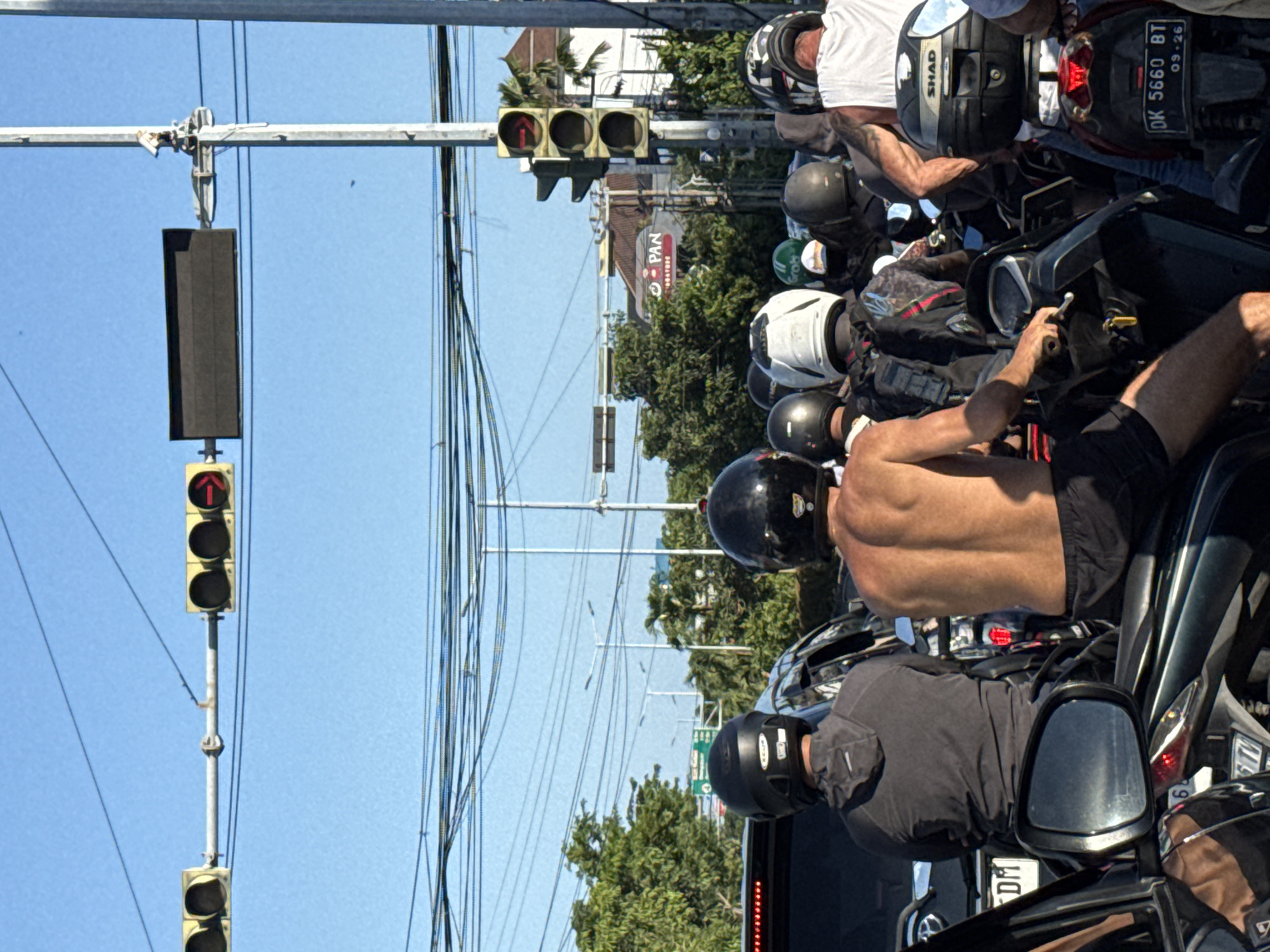 The image shows a busy street scene in Denpasar, Indonesia. A row of motorcycles is stopped at a traffic light that is red. Many of the motorcycle riders are wearing helmets; one rider is shirtless. A building with the word "PAN" is visible behind the motorcycles. Overhead, there are many power lines. The sky is blue.