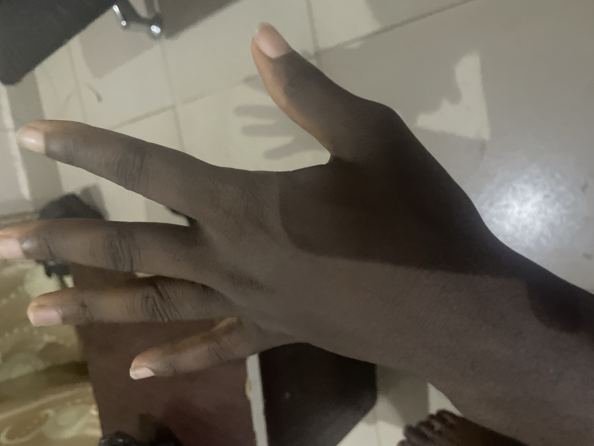 The image shows a close-up view of a dark-skinned hand, palm facing upwards, with fingers spread apart in a V-shape. The hand is positioned on a tiled floor, with a shadow cast on the floor that vaguely resembles another hand. A portion of a dark-colored wooden surface, possibly a table or desk, is visible in the upper left corner, and in the far left of the image, a part of a foot with visible toes can be seen. The lighting suggests an indoor environment, possibly a domestic setting. The image does not contain any specific visual cues that would allow for the identification of its location within the city of Jalingo, Nigeria.