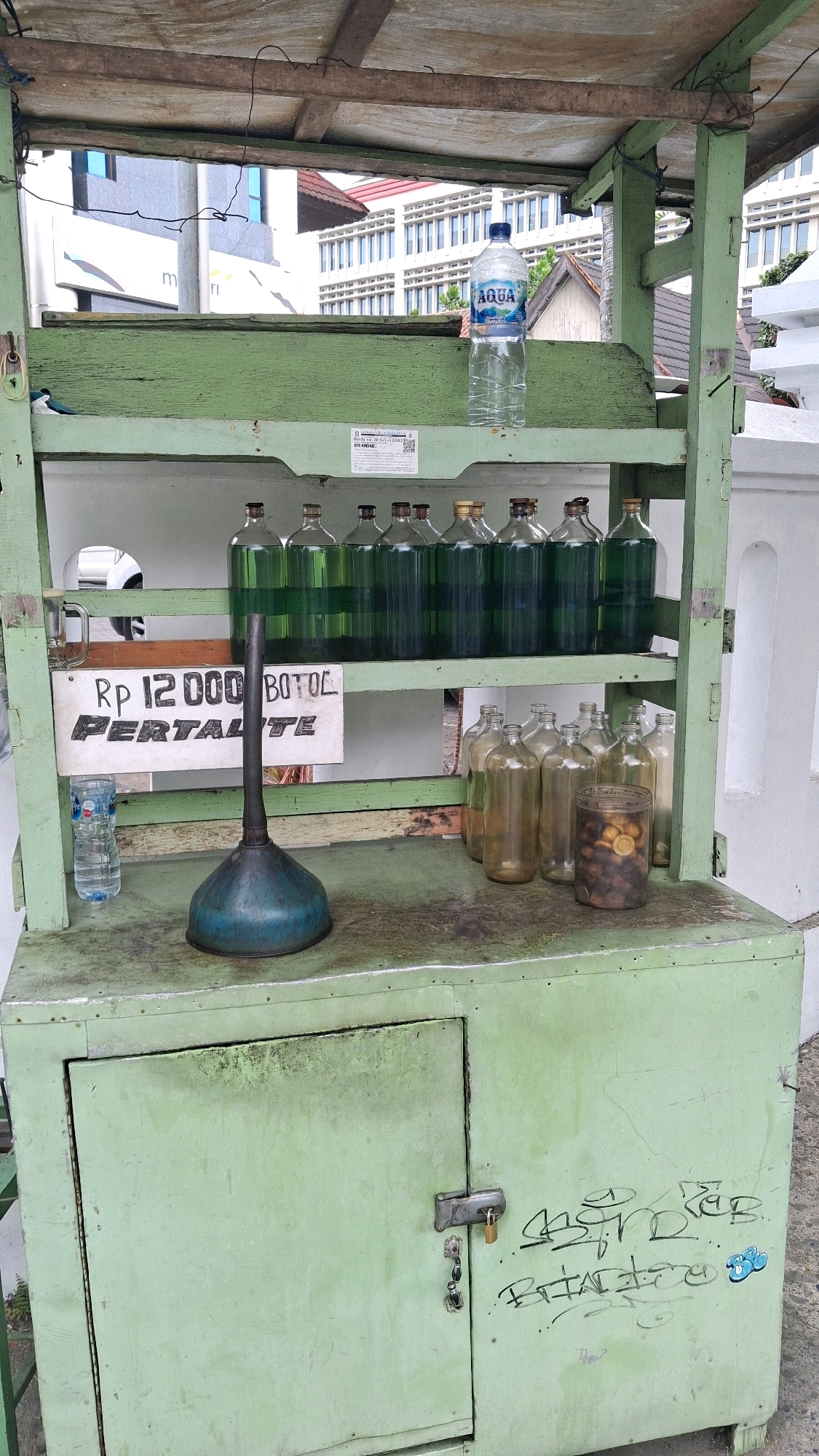 This is a medium shot, outdoor, eye-level, full-frame photograph of a roadside fuel stand in Yogyakarta, Indonesia. The stand is constructed from weathered, light green painted wood and metal. At the top, a sloped wooden roof is supported by vertical wooden posts. A wire hangs from the roof.
The stand has two shelves. The upper shelf holds a large plastic bottle of Aqua brand bottled water. Below that, the shelf displays rows of green glass bottles, each filled with a clear, green liquid, likely fuel. A sign is attached to this shelf, with black text on a white background. The text reads "RP 12 000 BOTOL PERTALITE". In front of the sign, a blue metal funnel with a long spout rests on the lower shelf.
To the right of the funnel, on the lower shelf, are several clear glass bottles, some empty and some containing a small number of brown objects and what appears to be a golden coin. To the left of the funnel is a small plastic bottle of Aqua water.
The base of the stand is a light green wooden cabinet with a hinged door secured by a padlock and two keys. The cabinet is covered in graffiti, including scribbled words and a cartoonish blue sticker. The ground in front of the stand is concrete, and a portion of a white wall is visible on the right side of the frame. The background shows parts of buildings and some greenery.