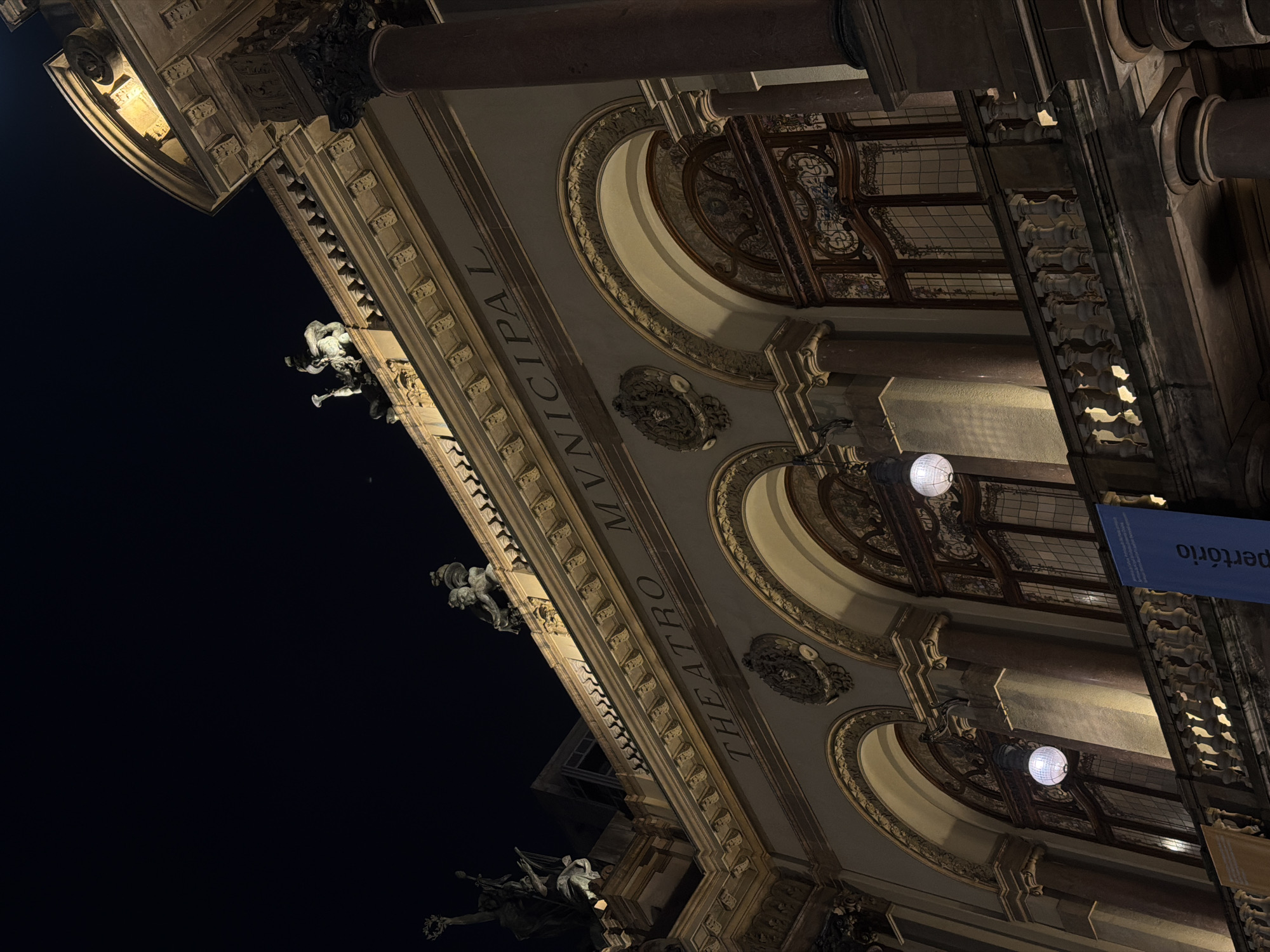 The image is a low-angle, nighttime exterior shot of the Theatro Municipal of São Paulo, Brazil. The facade of the grand building is illuminated, highlighting its ornate architectural details. Sculptures are visible on the rooftop, and decorative elements adorn the upper stories. Large arched windows with intricate wooden frames and stained glass are prominent. Below the windows, a stone balcony with a decorative railing is visible. Two spherical lights hang from the balcony, illuminating the facade. A blue banner with white text is hanging from the building, and part of a yellow banner is visible to the left. The sky above is dark and clear, indicating it is nighttime.