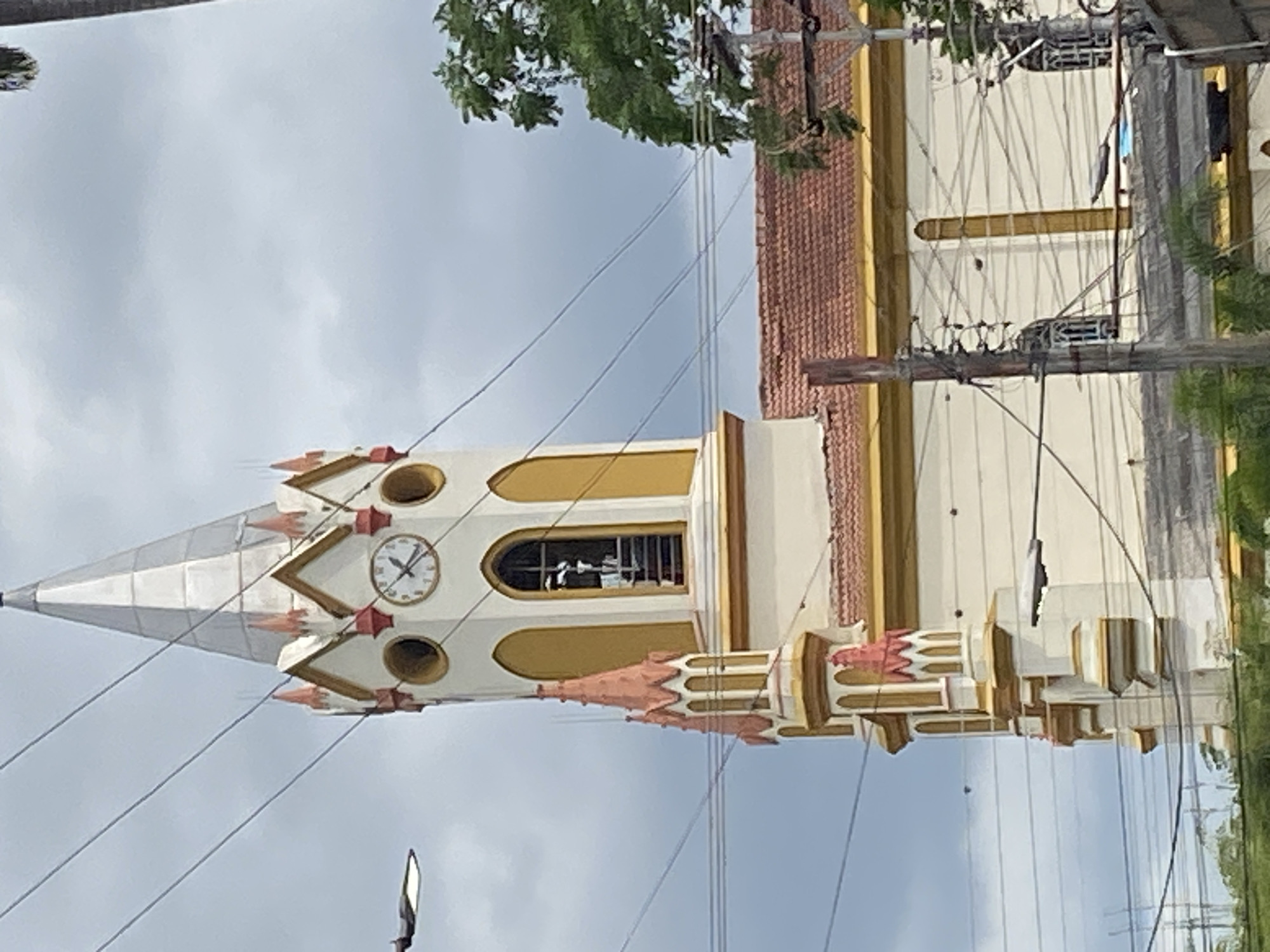 The image shows a vertical view of a church tower in Tinaquillo, Venezuela. The tower is painted in cream, white, and yellow hues, featuring a clock near the top. A white conical spire tops the tower. Power lines are visible in the foreground, crisscrossing the view of the tower. Overcast sky is visible in the background. The adjacent structure of the church, partially visible, has a brick-colored roof. Greenery suggests nearby trees.