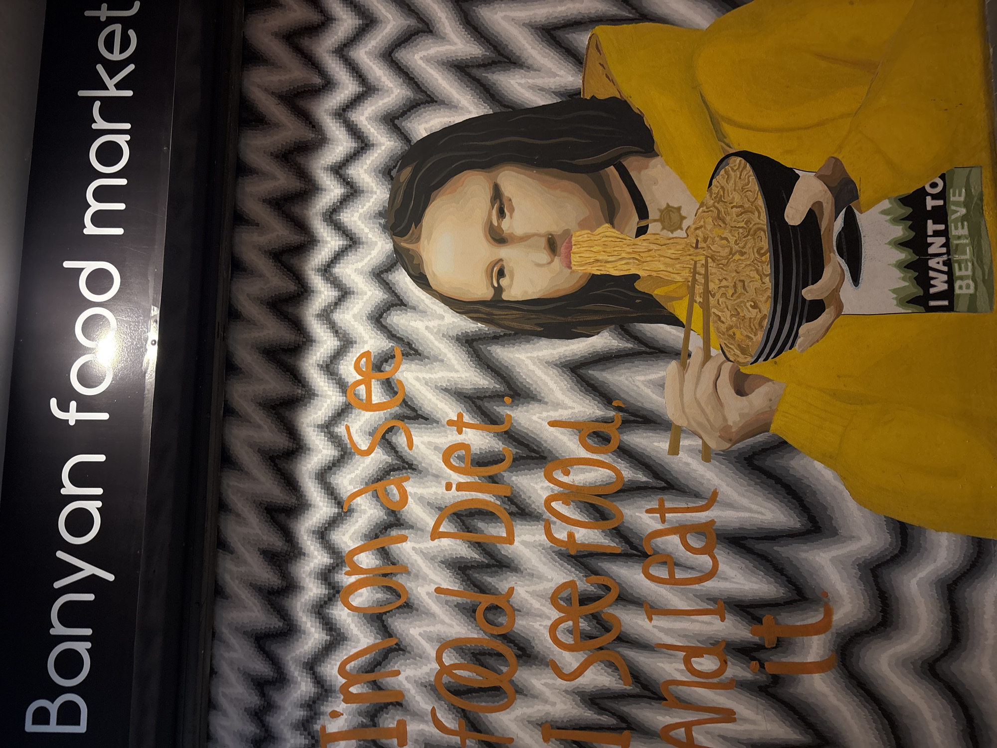 This image captures a vibrant mural or painting, likely located within or near the "Banyan food market" as indicated by the white text on a black panel to the left. The location is specified as บ้านบางรักษ์, Thailand.
The central subject of the artwork is a stylized person, possibly female, with long dark hair, fair skin, and wearing a yellow sweater. She is depicted engrossed in eating noodles from a black bowl with chopsticks, with strands of noodles extending from the bowl towards her mouth. She wears a black choker with a star-like pendant.
The background of the painting features a striking grey and white zig-zag or chevron pattern. On the patterned wall, large orange script reads: "I'm on a Diet I see food And I eat it". Another notable detail is a small green patch near the bowl, which contains the white text "I WANT TO BELIEVE," a reference to The X-Files. The overall tone of the mural is humorous and playful, celebrating food consumption.