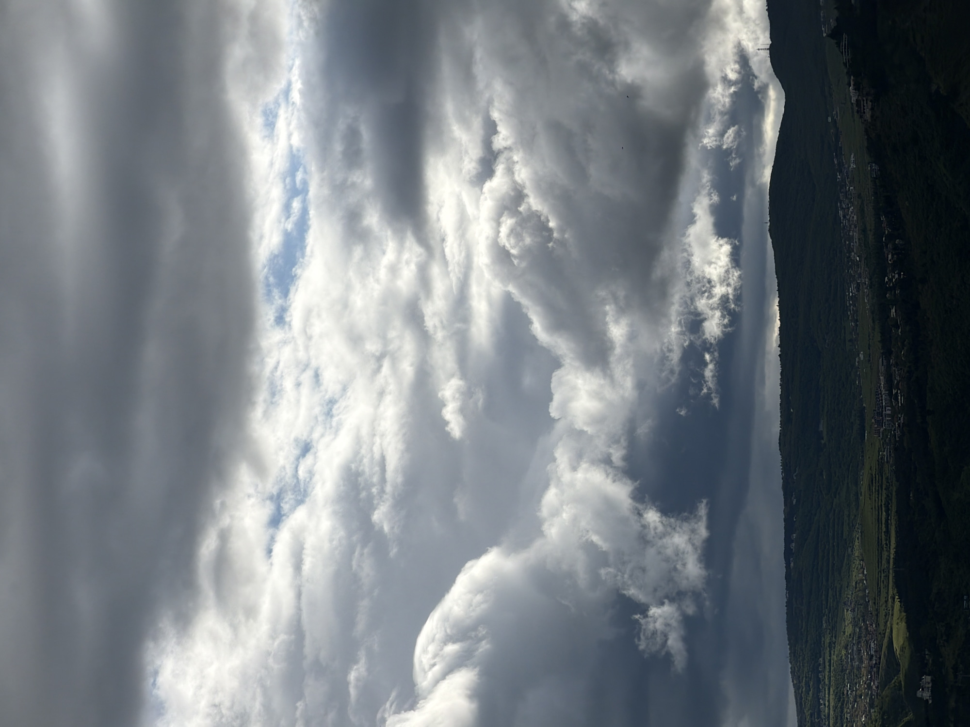The image displays a view of a mountain range from what appears to be a slightly elevated position. The sky is dominated by a variety of clouds, ranging from bright white cumulus clouds to darker, grey storm clouds. A patch of blue sky is visible between some of the clouds. On the right side of the image, a section of a mountain range is visible, covered in dense vegetation. Small buildings and structures can be seen dotting the lower slopes of the mountain. The location is likely Tbilisi, Georgia, given the characteristics of the mountainous terrain.