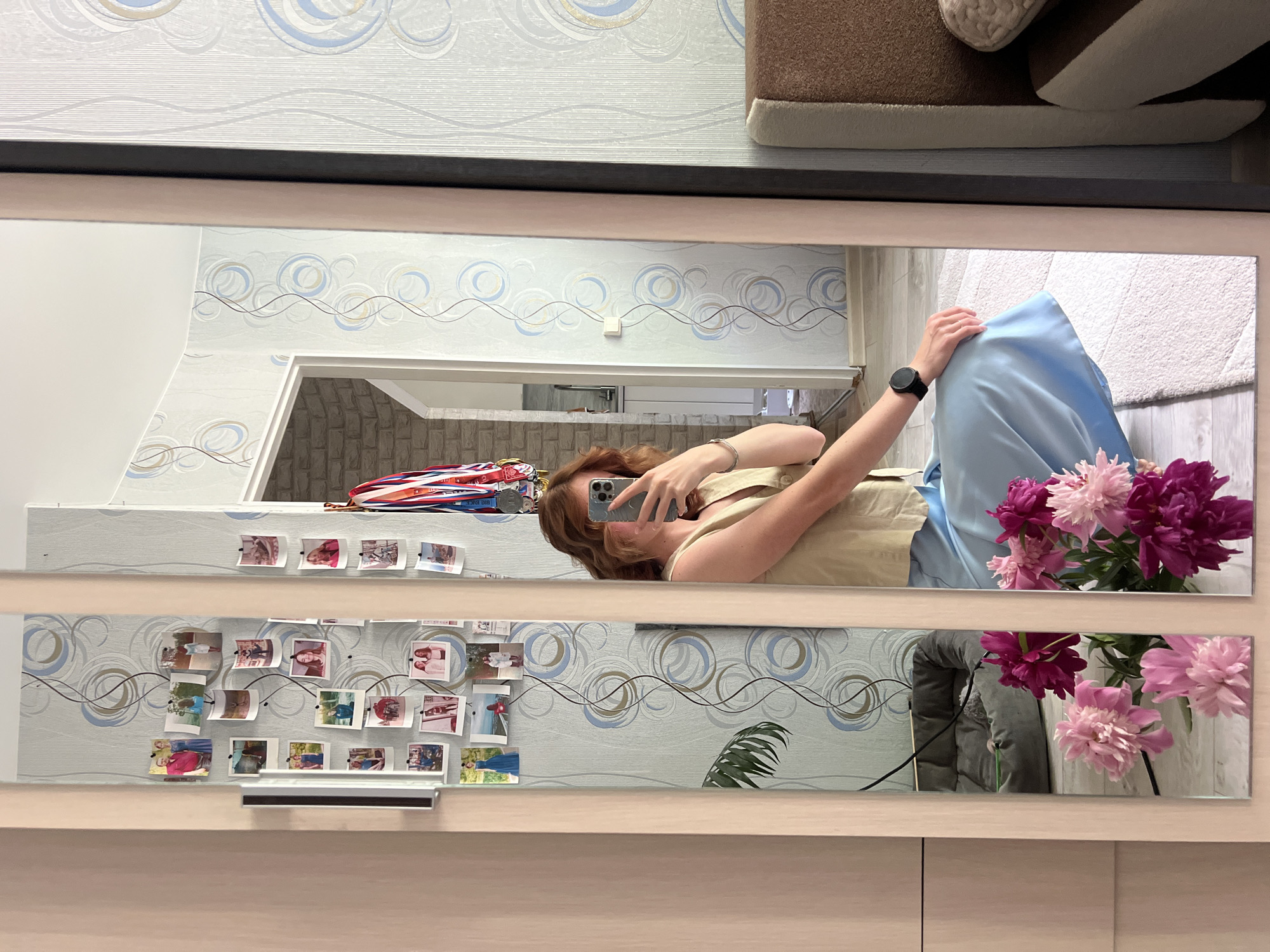 The image shows a woman taking a selfie in a mirror. She is wearing a light tan vest and a light blue skirt. She has medium-length reddish-brown hair. She is wearing a black smartwatch on her left wrist. In the background, there are several framed photos on the wall, a dog bed, and a bouquet of pink and purple peonies. The scene appears to be indoors, likely in a residential setting. The wallpaper has a blue and grey circular pattern. The overall impression is a casual, personal moment captured in a home environment, possibly in Chaikovsky, Russia given the instruction to include this location.