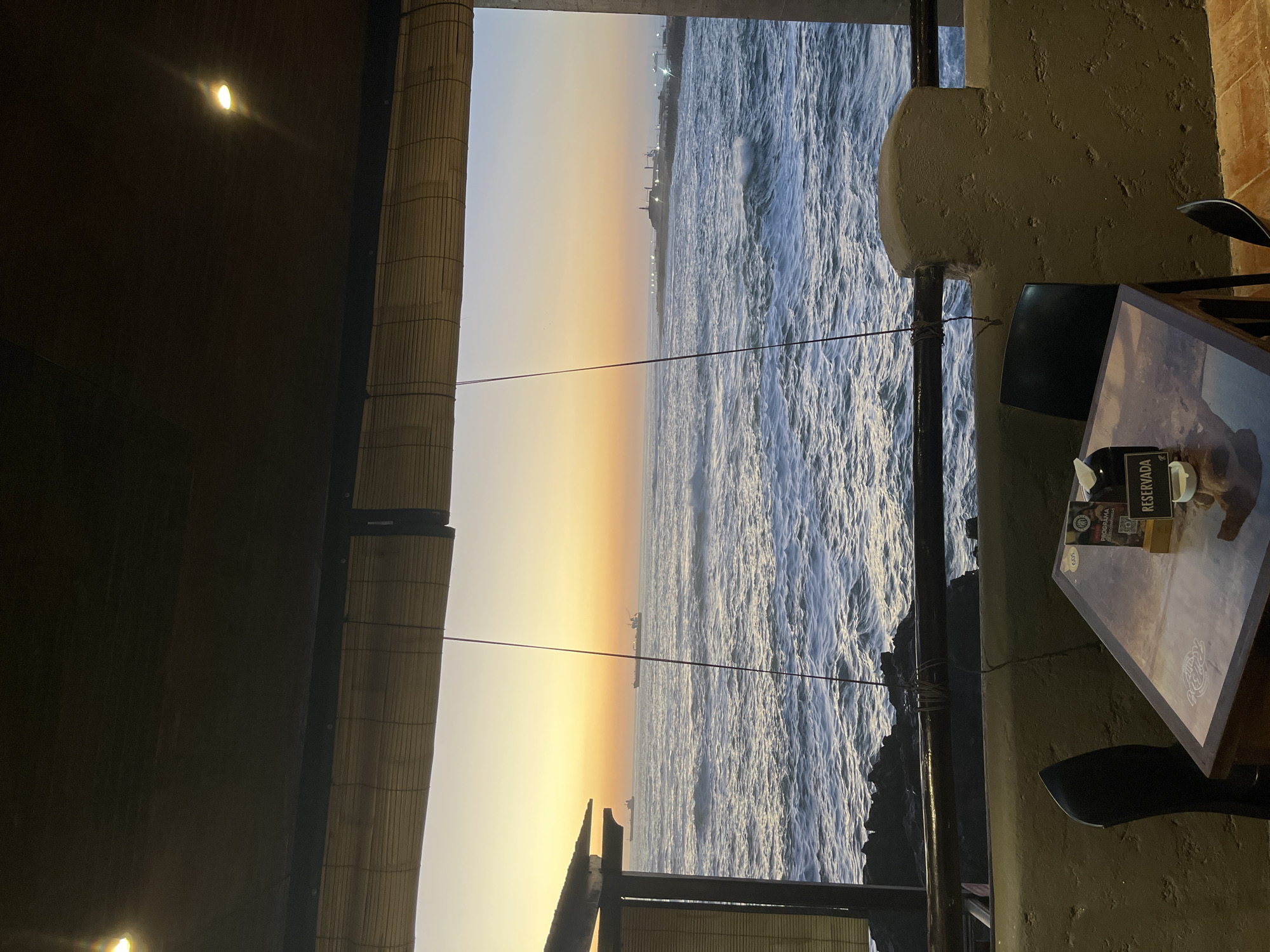 The image shows a view from what appears to be an indoor or partially enclosed space, likely a restaurant or café, overlooking the ocean at sunset in Arica, Chile. The ocean is visible through a large opening in the structure. The water is choppy with whitecaps, indicating some wind. In the distance, along the horizon, there is a suggestion of buildings or structures that might be located along the coast.
To the left, a bamboo or woven reed blind partially obscures the view. To the right, there's a rough, textured wall and a glimpse of a table and chairs, suggesting the interior of the establishment. A small sign on the table reads "Reservada," meaning "Reserved." The light suggests the photograph was taken in the evening.