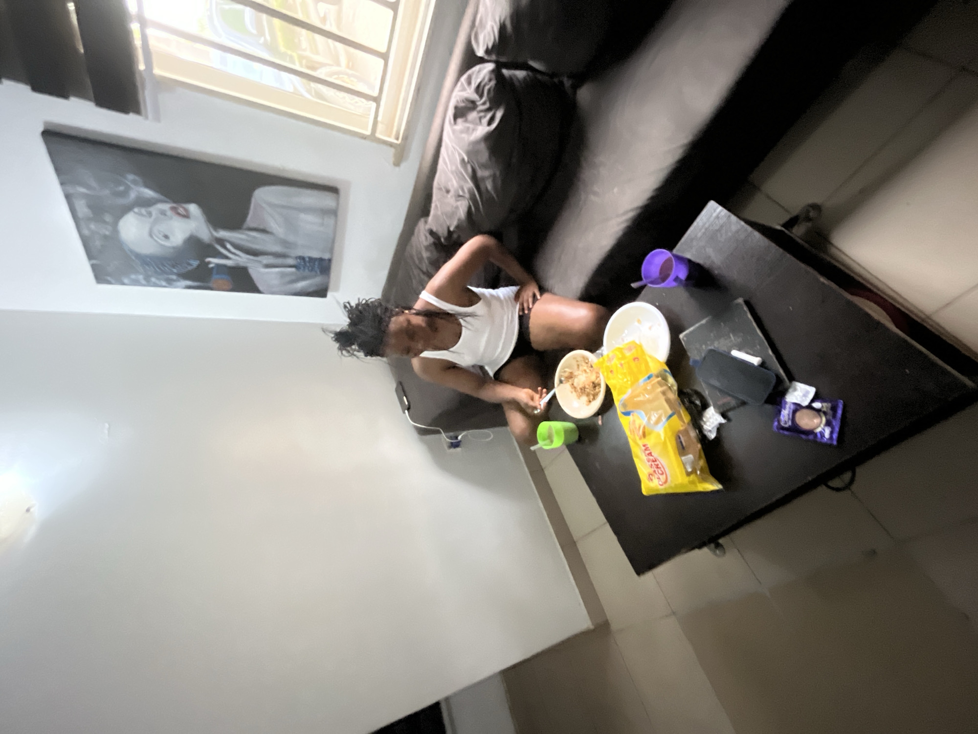 A young Black woman with braided hair, wearing a white tank top and black shorts, sits on a dark brown sofa in a living room, eating from a bowl of food. A black coffee table in front of her holds a yellow package of cookies, a white bowl, a green cup, a purple cup, and a small dark purple package. A framed painting of a woman smoking is on the wall behind her, and a window with blinds is to the right, showing a blurred exterior view. The floor appears to be tiled. The overall impression is a casual indoor setting.