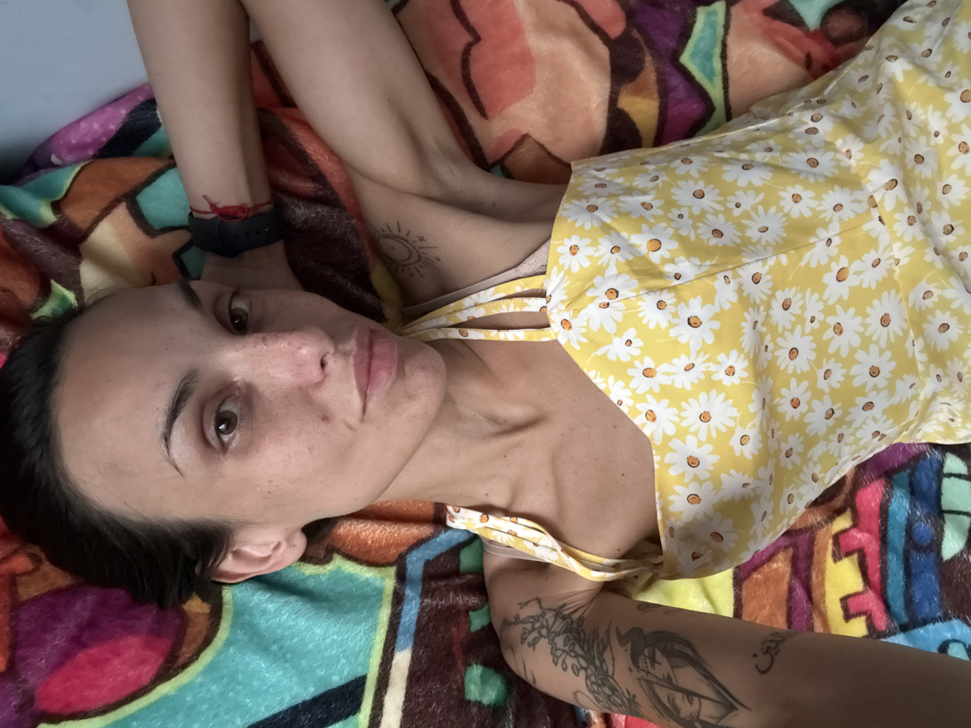 A young woman with dark hair and fair skin lies on her back on a colorful blanket. She is wearing a yellow dress with a daisy pattern. Her right arm is bent and her hand is resting behind her head. She has tattoos on her left arm and a small sun tattoo on her right shoulder. A red string bracelet is tied around her left wrist. The blanket has a geometric pattern with bright colors, including pink, orange, yellow, blue, and green. The image was taken in the city of Pleven, Bulgaria.
