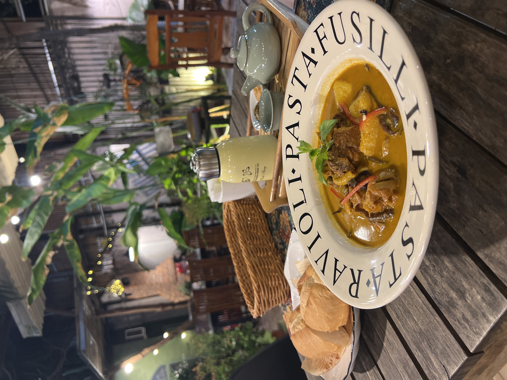The image shows an outdoor dining scene. A large bowl of yellow curry, garnished with basil, sits on a wooden table. The bowl is part of a dish set with "PASTA FUSILLI, RAVIOLI" printed around the rim. A plate of bread is next to the bowl. In the background, a light-green water bottle and a ceramic tea set sit on a serving tray. A woven basket sits to the side. The background features lush greenery and glimpses of a building structure with string lights. The setting appears to be an outdoor restaurant or patio. Nha Trang, Vietnam is inferred from the water bottle branding.
