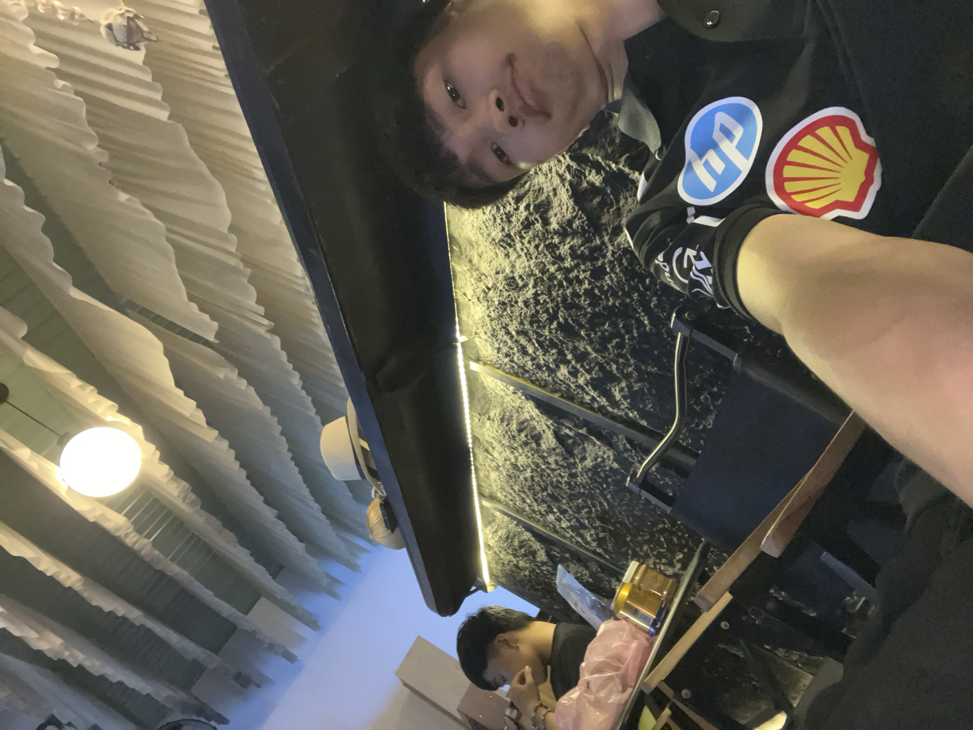 This indoor image, taken in Đà Nẵng, Vietnam, captures a selfie perspective from a young man. The main subject, a male with dark hair, looks directly at the camera with a slight smile. He is wearing a black long-sleeved top that prominently displays two circular logos on the chest: a blue one with "hp" (Hewlett-Packard) and a red and yellow scallop shell (Shell). His right forearm is visible in the foreground.
The setting appears to be a cafe or restaurant, indicated by the interior decor and a second individual. Behind the main subject, a dark, textured wall, possibly stone or concrete, is illuminated by a warm strip of LED lighting. In the mid-ground, another young man with dark hair is seated at a table, looking down, engaged in an activity that isn't fully visible. On the table near him, a golden container and a pink plastic bag can be seen.
The ceiling features an intricate design with light-colored, wavy panels or fabric and a bright circular light fixture. A light-colored hat hangs on a nearby structure. The lighting suggests it is evening or night. The overall atmosphere is casual and relaxed.