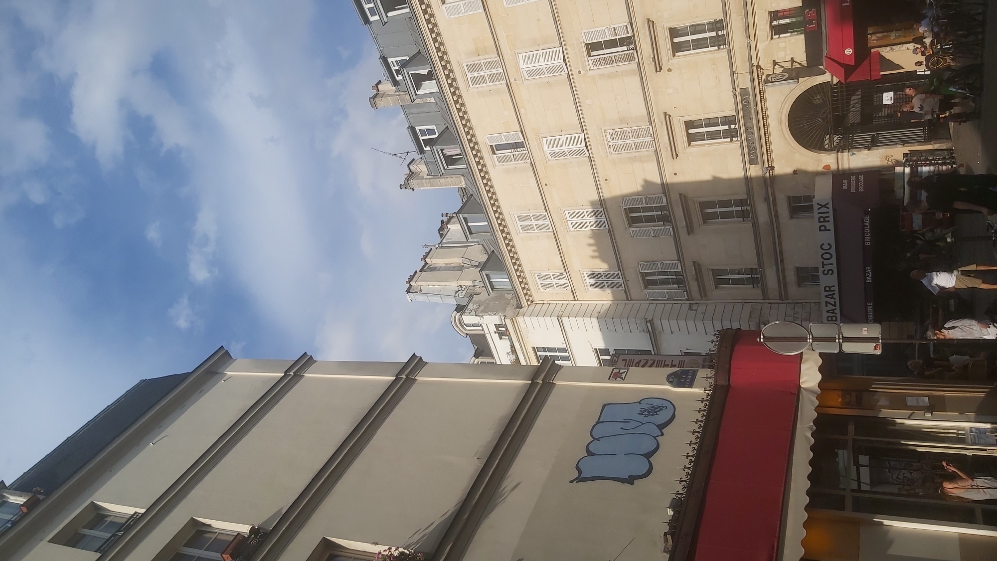 The image captures a street scene in Paris, France, likely in the afternoon, with buildings bathed in sunlight and some shadowed areas. In the foreground, to the left, a cream-colored building with dark gray mansard roof dominates the frame. A section of this building features a large graffiti tag in blue. Below this building, a storefront with a red awning and a white canopy is visible, with people partially obscured inside. A street sign and a traffic sign are also present.
To the right, a taller, light-yellow building with multiple floors of shuttered windows is prominent. Above an arched entryway, the text "PASSAGE D'EUX" is inscribed. This building appears to be residential or commercial. Further to the right, another building with a red awning is visible, and people are gathered outside, some seated at tables and others standing. The street is lined with bicycles. The sky above is blue with scattered white clouds. The overall impression is of a bustling urban environment in Paris.