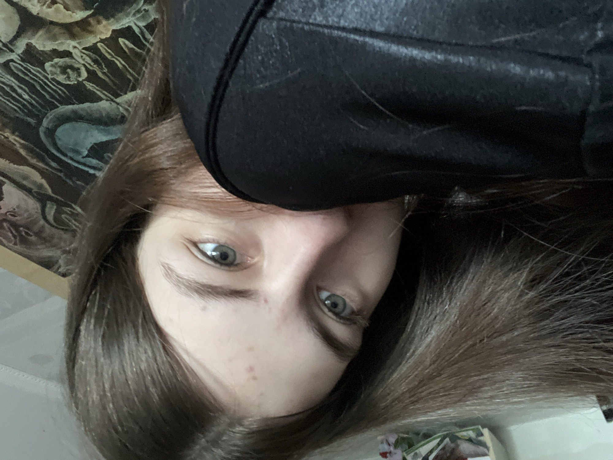 The media file presents an inverted, close-up portrait of a young person, likely female, with long, straight brown hair and light blue or green eyes. Her face is positioned upside down, with her eyes looking directly at the viewer. She has a neutral expression, and a few small blemishes are visible on her forehead and nose. She is wearing a black garment, possibly a jacket or hoodie, which is visible around her shoulder and upper chest.
The background on the left side features an ornate, dark-toned pattern, possibly a tapestry, wallpaper, or painting, showcasing abstract, wavy shapes in muted shades of green, brown, and black. This pattern suggests an indoor setting. The right side of the background is less defined but also indicates an interior space, with some indistinct, lighter elements at the bottom. The lighting is soft and appears to be even, consistent with an indoor environment. There are no identifiable location cues beyond an interior setting, nor any visible text. The time of day is not discernible.