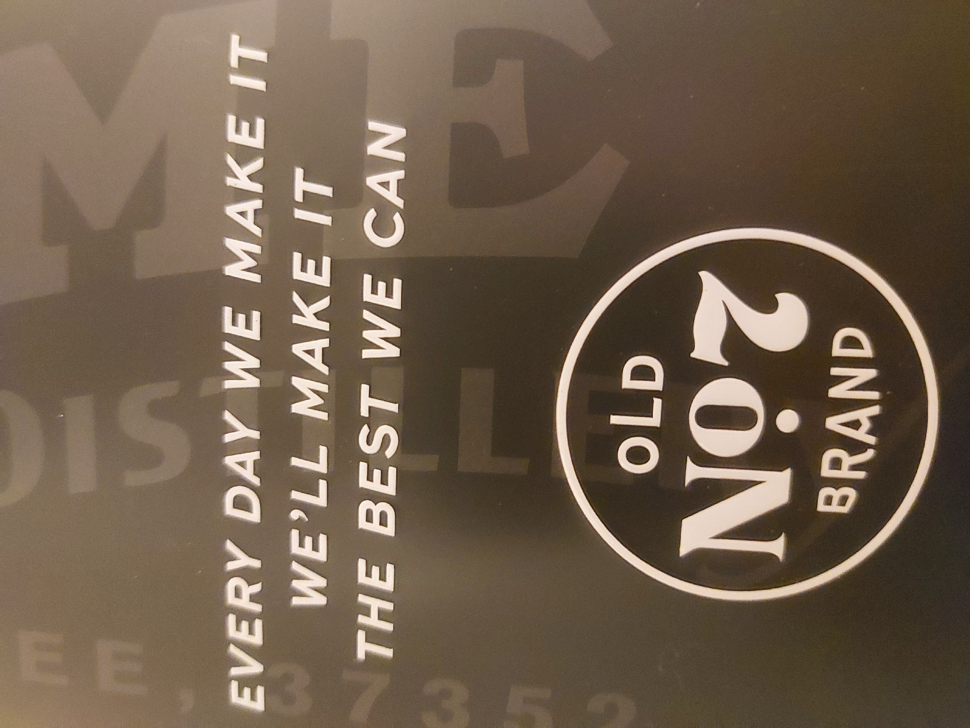 The image shows a black and white Jack Daniel's Old No. 7 Brand advertisement. Text to the left reads, "EVERY DAY WE MAKE IT, WE'LL MAKE IT THE BEST WE CAN." Other partially visible text includes "DIS..." and "EE, 37352." No people are visible. The location of the scene is not stated in the image.
