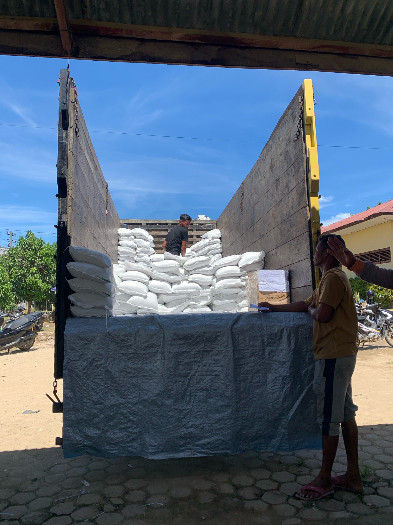 In Pantegurah, Indonesia, a flatbed truck is parked, its wooden sides raised, and the bed covered with a grey tarp. The truck is laden with numerous white sacks, stacked high, presumably containing goods. Two men are visible. One man, wearing a black t-shirt, stands amidst the sacks in the truck bed. Another man, in a light brown t-shirt and grey and blue striped shorts, stands to the right, facing away from the camera, with another person's hand resting on his head. A cardboard box with some writing on it, including "MINYAKITA," sits on the truck bed near the second man. In the background, a building with a red roof and a yellow wall is visible, along with trees and parked motorcycles. The sky is bright blue with scattered clouds, indicating daytime.
