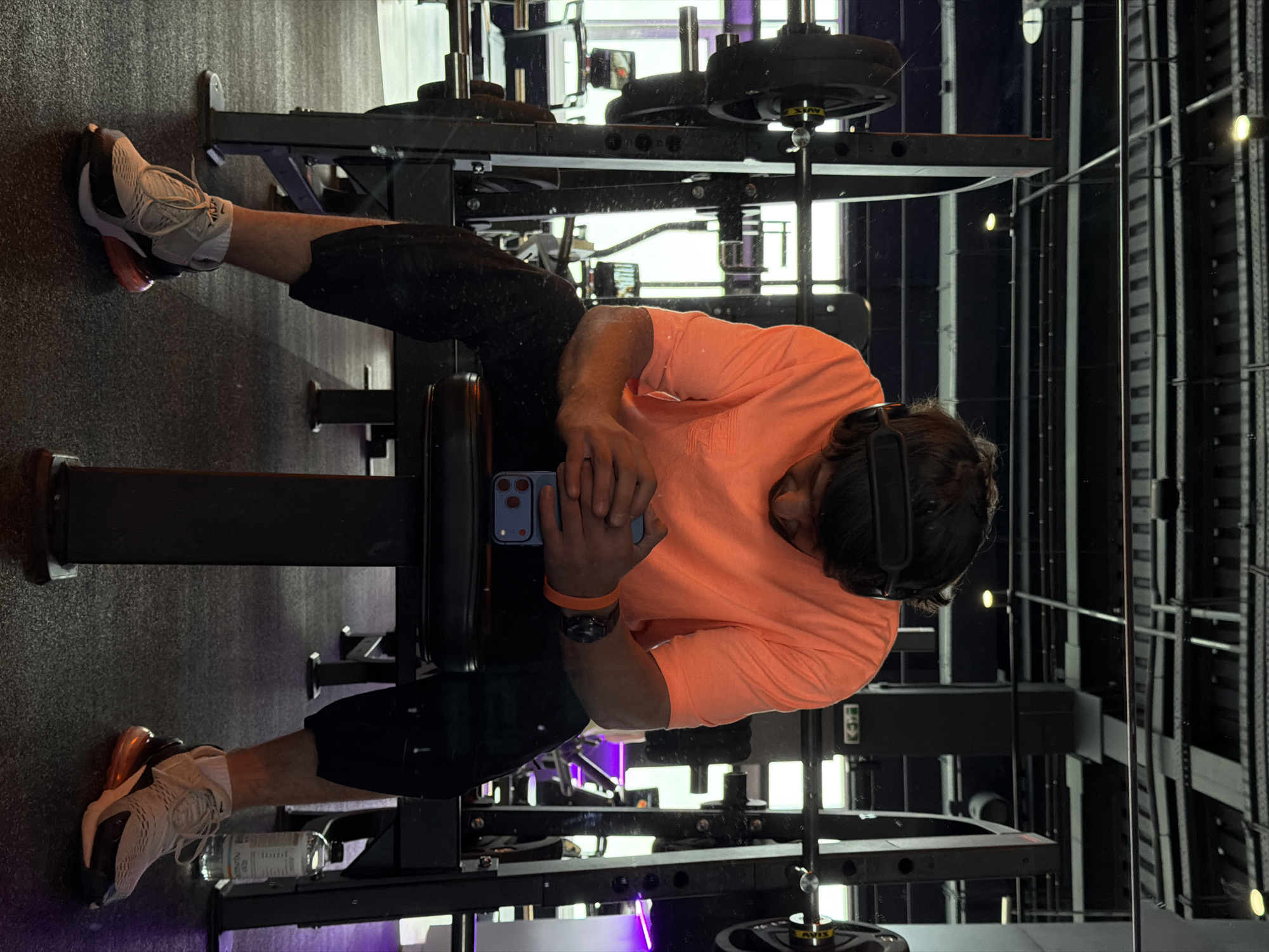 A man is sitting on a bench in a gym, looking at his phone. He is wearing a salmon-colored t-shirt, black shorts, and sneakers. He has a watch on his left wrist and is holding a blue iPhone in his hands. He is wearing a VR headset on his head. The gym has weightlifting equipment, including a squat rack with weights on it. The walls are black with exposed metal framework. There is a purple light strip along the bottom edge of the frame. The location and the city are not discernible from the image.