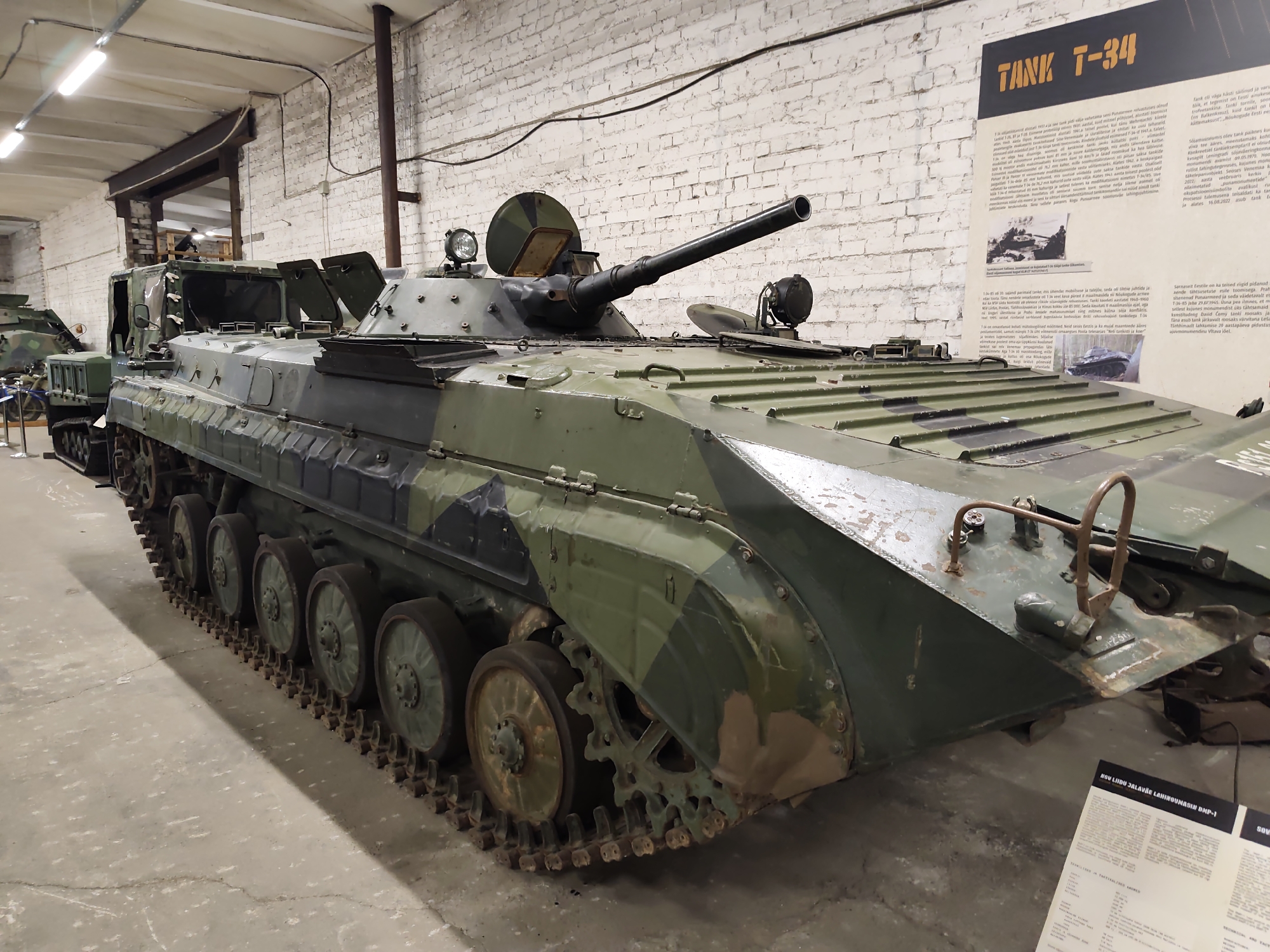 A dark green and black camouflaged tracked military vehicle, likely an infantry fighting vehicle, is displayed indoors on a concrete floor. The vehicle is positioned diagonally, facing towards the right of the frame. It features a prominent cannon extending from the turret. The vehicle is heavily built with visible suspension components and tracks. In the background, a white brick wall is illuminated by fluorescent lighting. A large informational poster titled "TANK T-34" is mounted on the wall to the right of the vehicle, featuring text and images related to a T-34 tank. Another smaller informational sign is visible on the floor in the lower right corner of the image. The overall environment appears to be a museum or exhibition space. The vehicle is identified as an "ASU LIHDU JALAVAE LAHMMIGUNA BMP-1" on the smaller sign on the floor, and further information about a T-34 tank is on the larger poster, suggesting the museum may be related to military history. This scene is located in the Viimsi Open Air Museum (Viimsi vabaõhumuuseum) in Viimsi, Estonia.