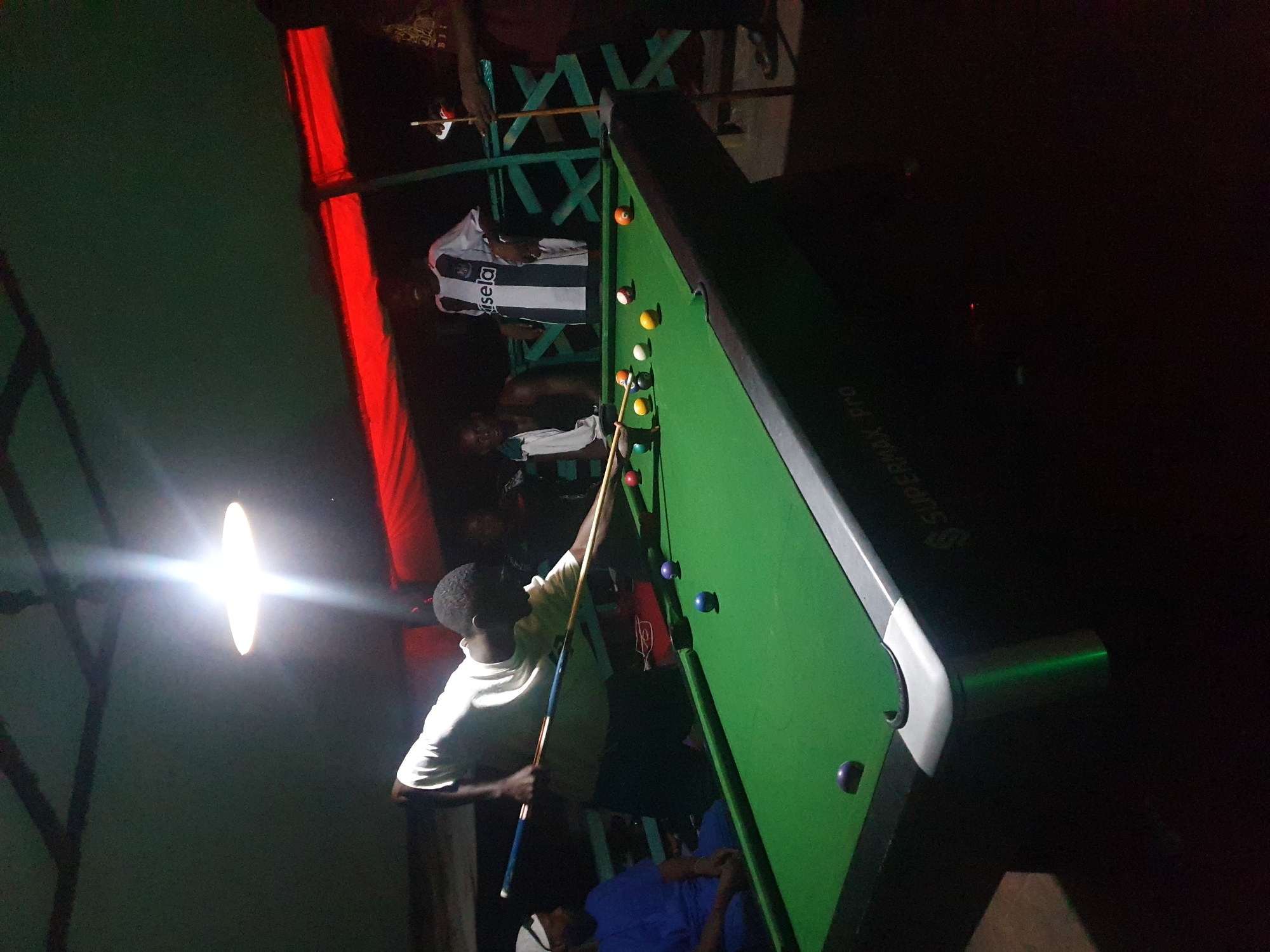 A nighttime exterior scene at what appears to be an outdoor recreational facility in Lokoja, Nigeria, features a group of individuals engaged in a game of pool. A bright overhead light illuminates the green pool table, upon which several colored billiard balls are visible. A man in a white t-shirt is actively playing, aiming his cue stick. Another man in a blue shirt sits to the left, observing the game. In the background, other individuals are present, some standing near a structure covered by a red awning, suggesting a social gathering or entertainment venue. The overall atmosphere is casual and social, set against a dark, open-air environment.