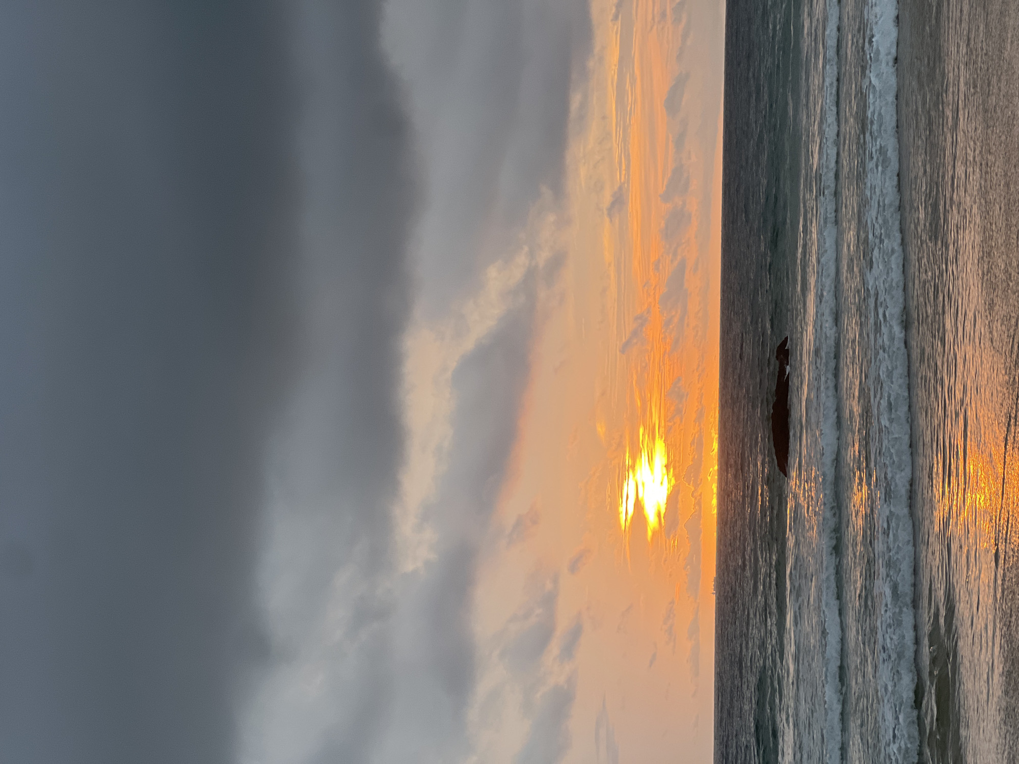 An ocean sunset is captured from a beach in Choeng Thale, Thailand. The sky is a dramatic mix of dark, heavy clouds towards the top and a vibrant orange and yellow hue from the setting sun closer to the horizon. The sun itself is a bright, intense glow peeking through the clouds. Below the sky, the dark ocean stretches out, with gentle waves rolling towards the sandy shore. A solitary dark rock formation is visible in the water, and in the far distance, on the horizon, a few distant ships or structures can be discerned. The wet sand at the edge of the water reflects the warm colors of the sunset. There are no people or animals visible in the frame.