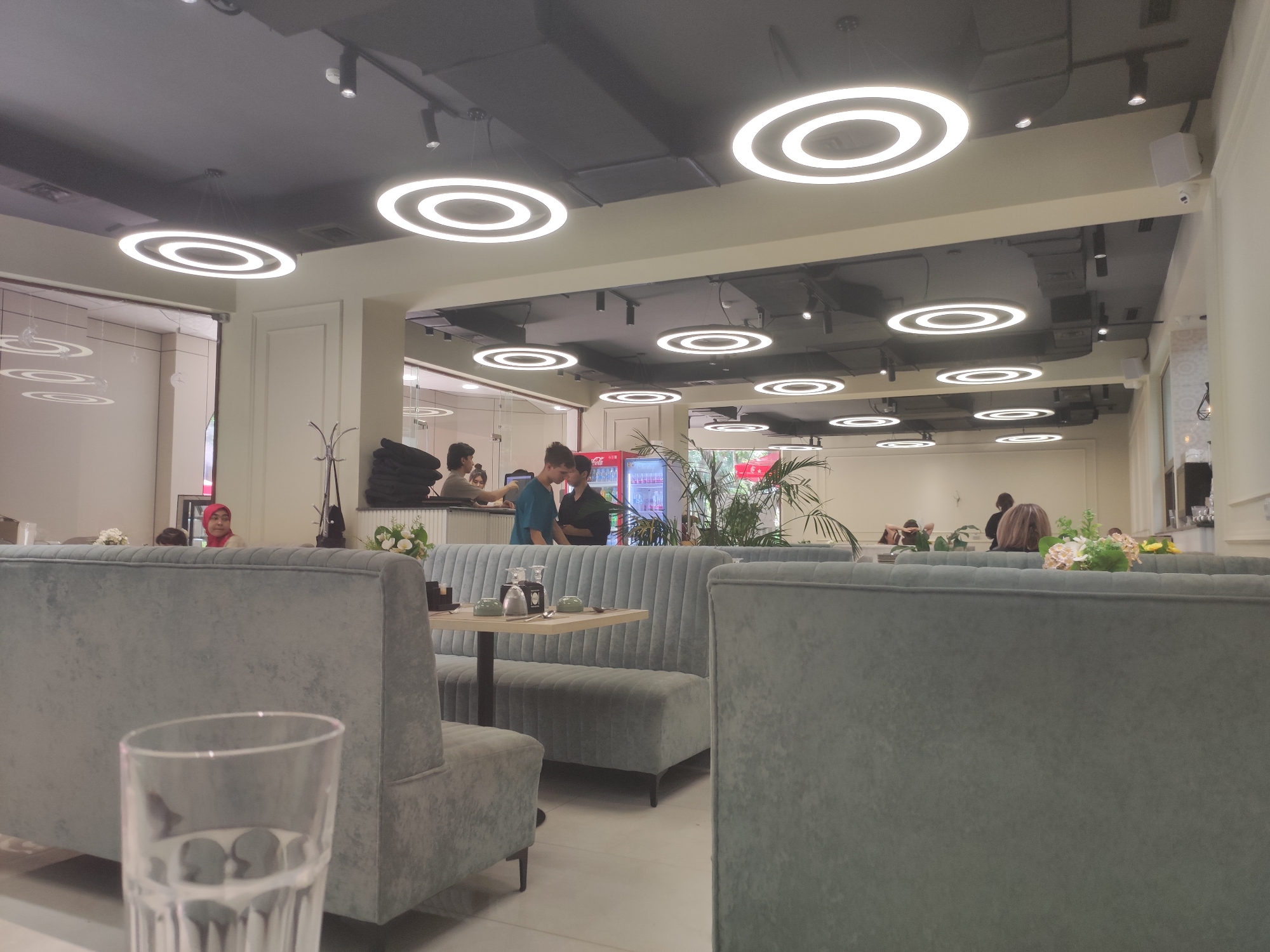 The image depicts the interior of a modern, upscale restaurant with a minimalist design. The ceiling features numerous circular, illuminated light fixtures. The seating consists of plush, light blue upholstered booths and chairs. Several tables are visible, some set with tableware, including glasses, soy sauce bottles, and small bowls. In the background, a red Coca-Cola refrigerator is visible, along with several potted plants. A few individuals are present in the scene: two young men are interacting at a table, one woman wearing a red hijab is seated at a booth, and other patrons are seated in the distance. The overall ambiance suggests a clean and contemporary dining establishment.