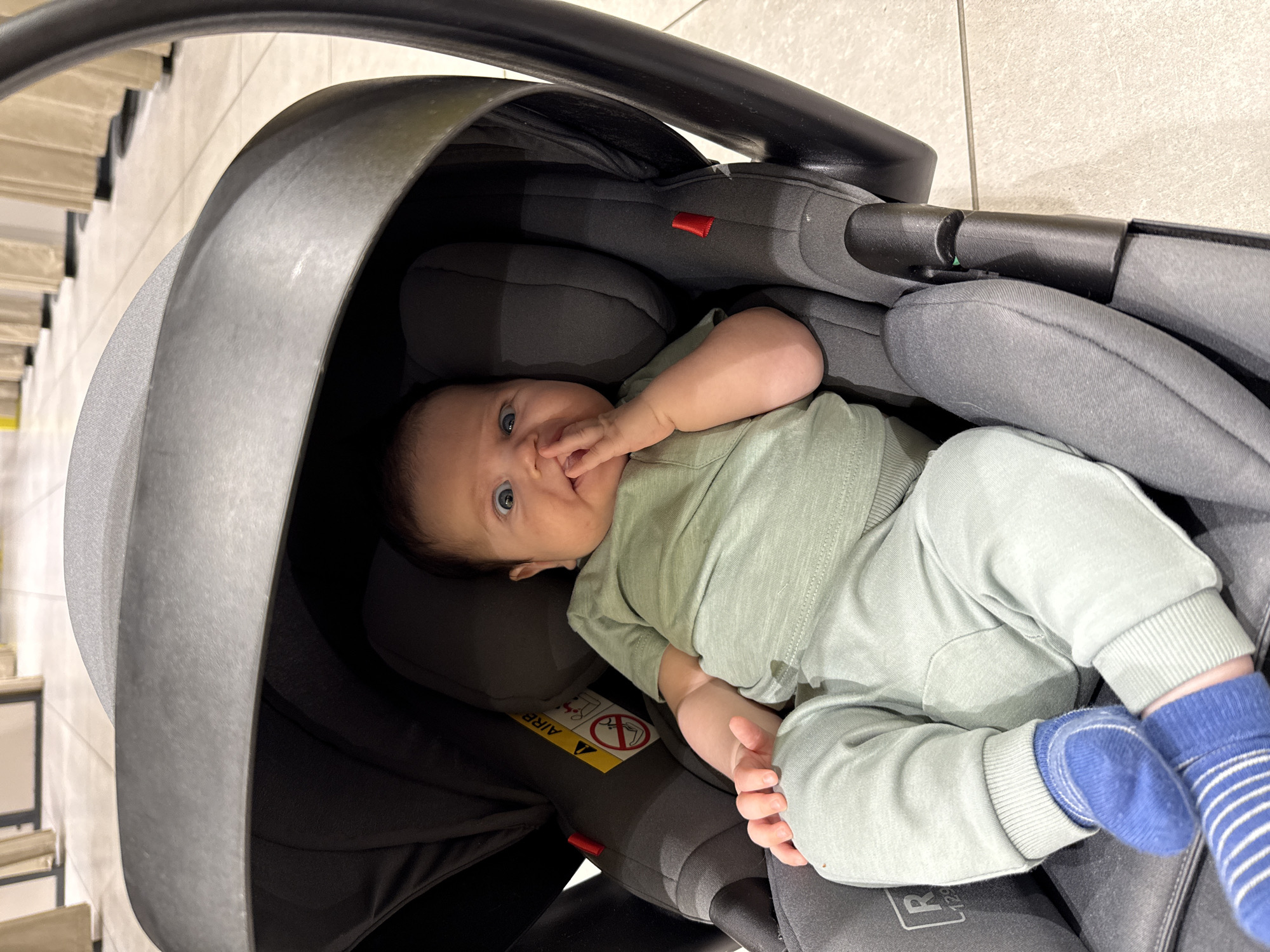 A close-up, overhead shot captures a baby lying in a car seat. The baby, with light skin and dark hair, is wearing a pale green outfit and blue and white striped socks. Its right hand is in its mouth, and its blue eyes are looking directly at the camera. The car seat is predominantly black with grey padding and a grey handle. A red warning label with symbols is visible on the car seat padding. The background consists of a tiled floor and a section of what appears to be a staircase with light-colored steps and dark risers. The lighting suggests an indoor setting, possibly during the daytime. The overall mood is calm and observational, with the baby appearing alert and engaged. No specific time of day or weather conditions can be determined from the image. Text visible on the car seat includes "AIRB" and "R 129".