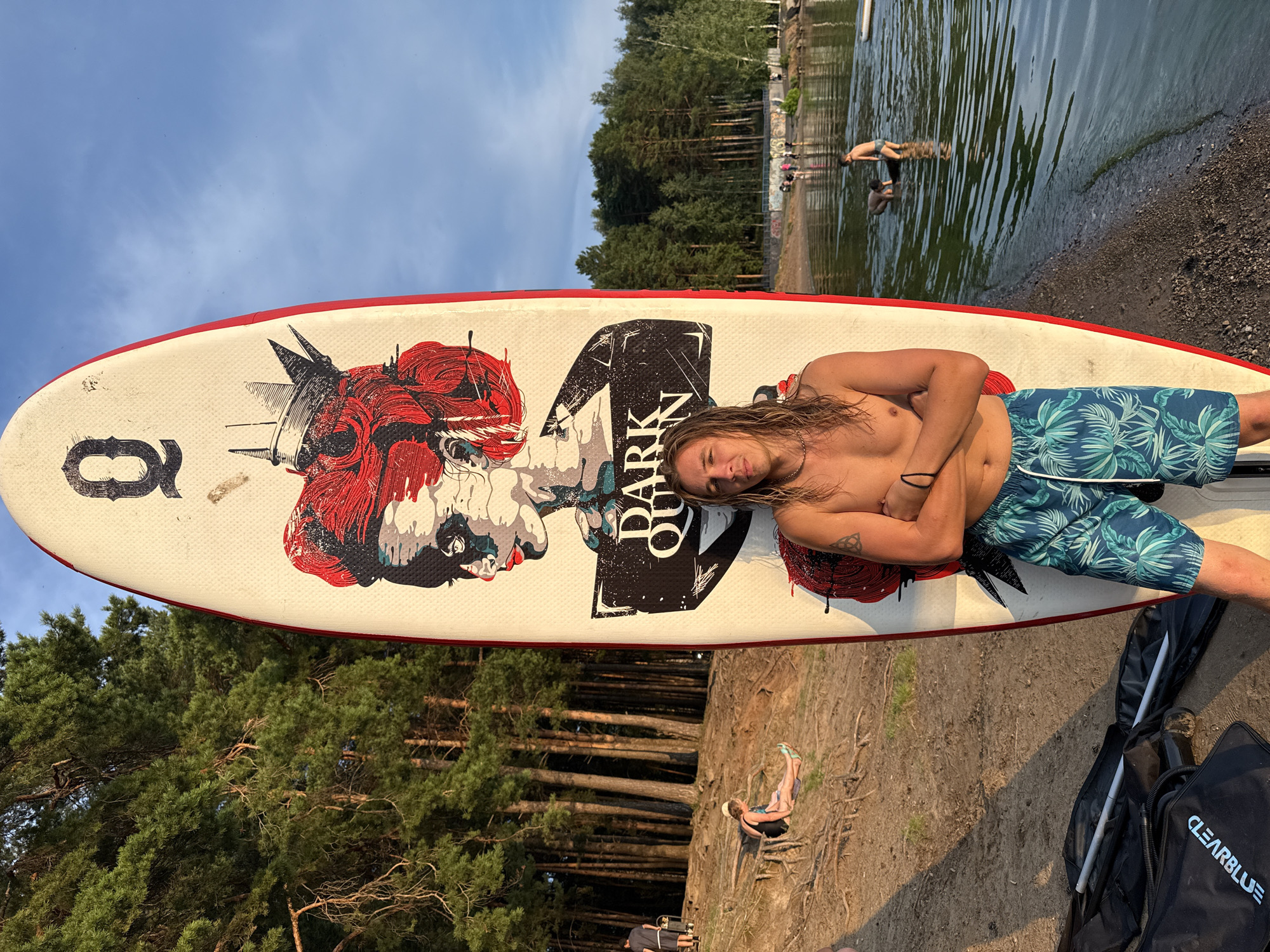 A young man with long blond hair and a tattoo on his left arm stands with his arms crossed in front of a paddleboard on a sandy beach. He is wearing patterned swim trunks. The paddleboard has a graphic of a stylized woman's face with red hair and a crown, and the words "DARK QUEEN" printed on it. Behind the man, a dense forest of pine trees lines the shore of a lake or river. Several other people are visible in the background, some near the water and others on the beach. The sun appears to be setting, casting a warm glow on the scene.