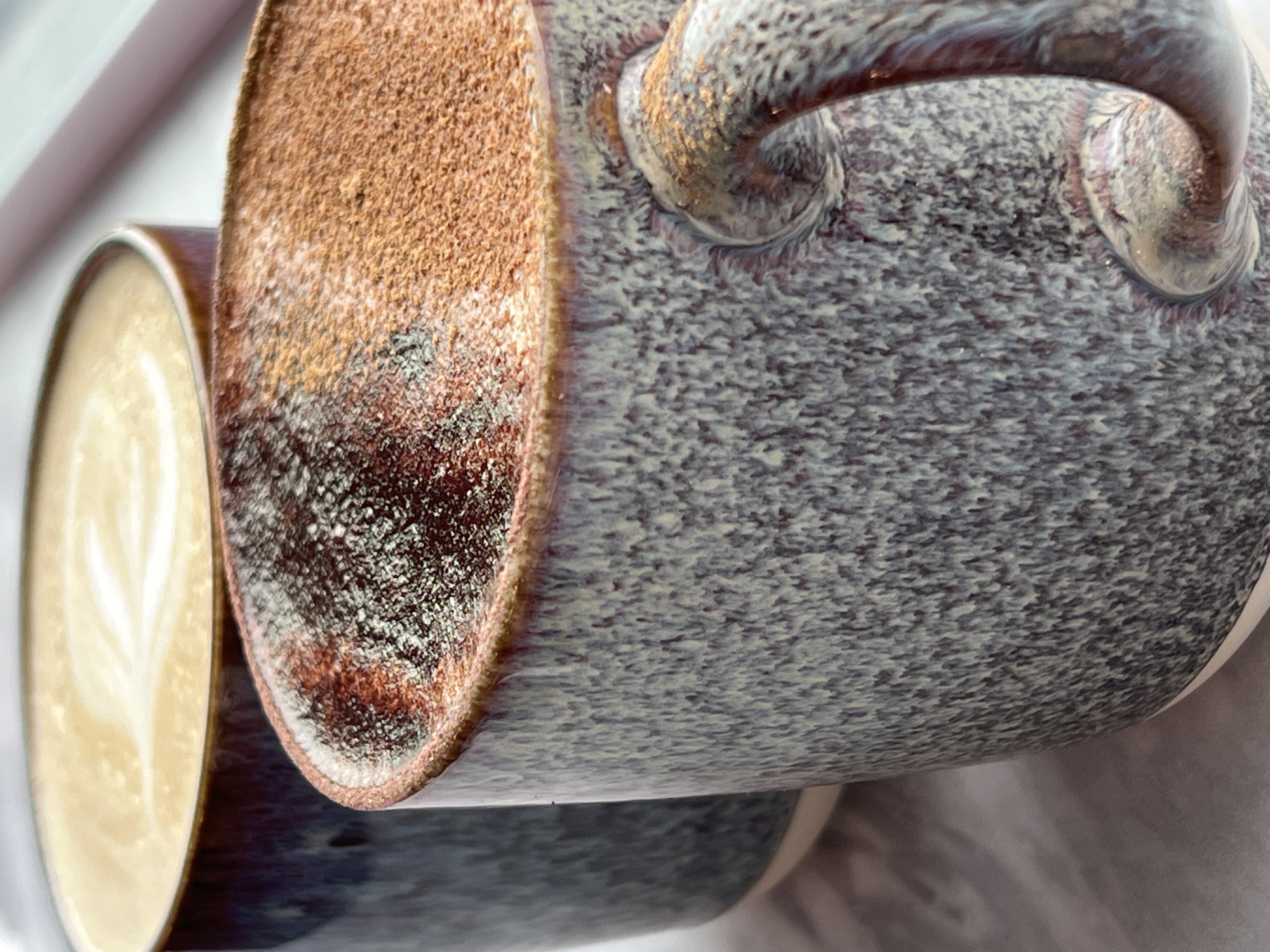 The image displays two mugs of coffee. One mug, in the foreground and slightly to the right, is partially filled with coffee dusted with cinnamon and has a textured, speckled glaze in shades of grey, blue, and purple, with a prominent handle. The second mug is positioned behind and to the left, filled with a lighter colored coffee featuring latte art, and has a darker, more uniform glaze. The mugs are set against a neutral background, with a hint of a window or ledge in the upper right corner. This scene is likely situated in a cafe or home setting in the city of Donegal, Ireland, given the request's context.
