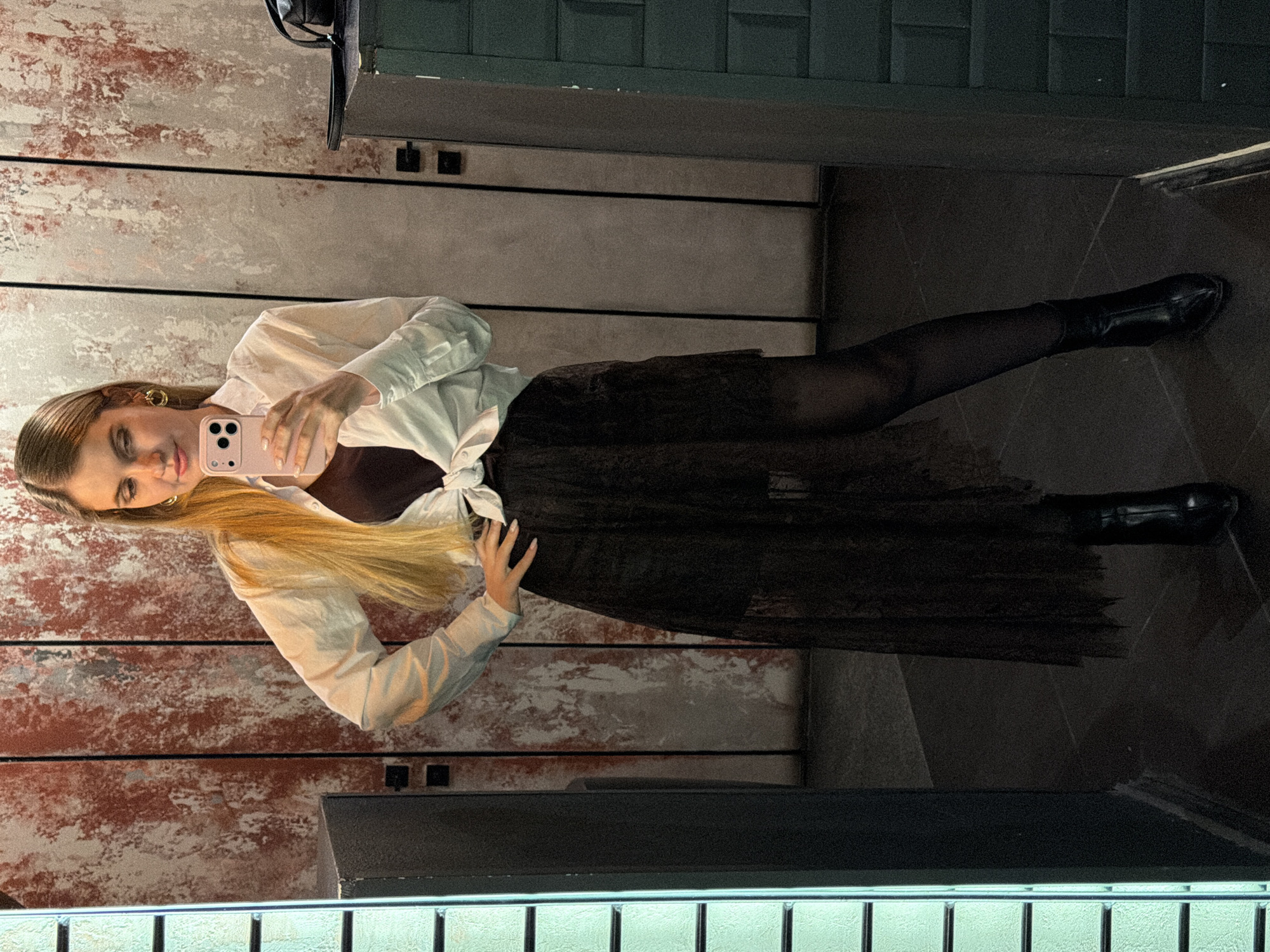 A woman in a white long-sleeved shirt, a dark brown top, and a sheer black skirt poses in a mirror selfie, likely in a bathroom. She has long blonde hair and is holding a pink phone. She is wearing black tights and black boots. The background features a textured wall with red and gray hues, a dark gray tiled wall, and a metal grate at the bottom of the mirror. This image could have been taken in Izhevsk, Russia, based on the general aesthetic and style of the environment, however, definitive confirmation would require more localized, specific details.