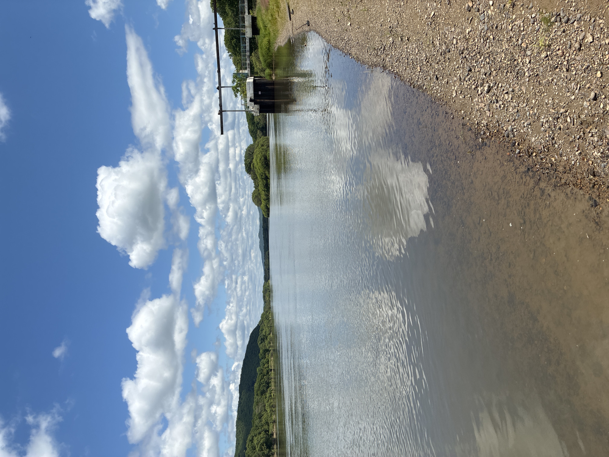 An overcast day is captured over a wide river that stretches into the distance, with the sun breaking through in the sky. A concrete structure with metal beams extends over the water from the right bank, likely a part of a dam or water control system. Lush green trees line the riverbanks, with hills in the background. The water's surface is calm, reflecting the sky and clouds, with a stony, sandy shore on the right. The location is Gornye Kljuchi, Russia.