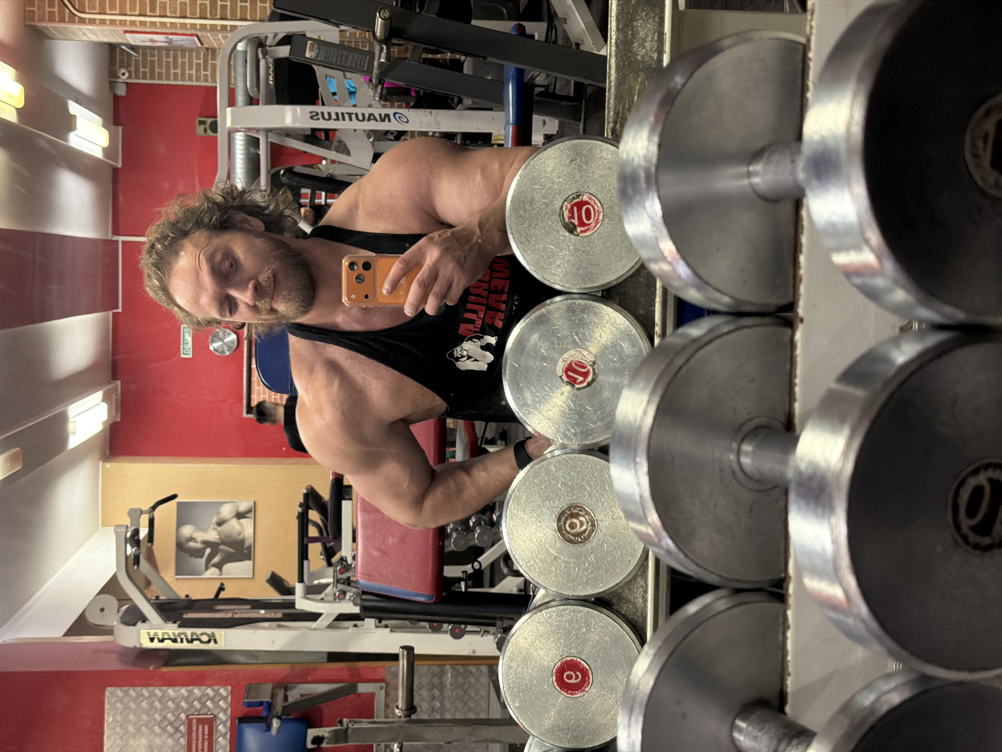 An indoor gym scene is depicted, with the primary subject, a muscular male with curly light brown hair and a beard, taking a mirror selfie. He wears a black tank top featuring a white gorilla logo and "GORILLA WEAR" text (reversed due to reflection) and a black fitness tracker on his left wrist. He holds an orange smartphone in his right hand, capturing his reflection. The subject's left arm is bent, hand resting on his hip, and he exhibits a slight smirk or wink.
In the immediate foreground, a rack of silver dumbbells is prominently reflected in the mirror. Several dumbbell plates are visible, displaying red or brown centers with markings such as "10" and a stylized "e".
The background, also visible through reflection, features various gym equipment including a lat pulldown machine bearing the Cyrillic brand "VARYAG" (ВАРАЯГ) and another machine with "NAUTILUS" branding. Walls are segmented into sections of red, orange-yellow, and a reddish-brown brick pattern. A framed image of a muscular man is affixed to the orange-yellow wall. Overhead, fluorescent tube lights illuminate the area. A white sign with red Cyrillic text "ВЫХОД" (Exit) on a green background is partially visible, confirming a Russian-speaking environment. A small speaker is mounted above the brick wall section. Another individual with short dark hair, wearing a black shirt, is partially visible in the reflected background.
The presence of the "VARYAG" equipment brand and the "ВЫХОД" sign firmly places this location in Saint Petersburg, Russia.