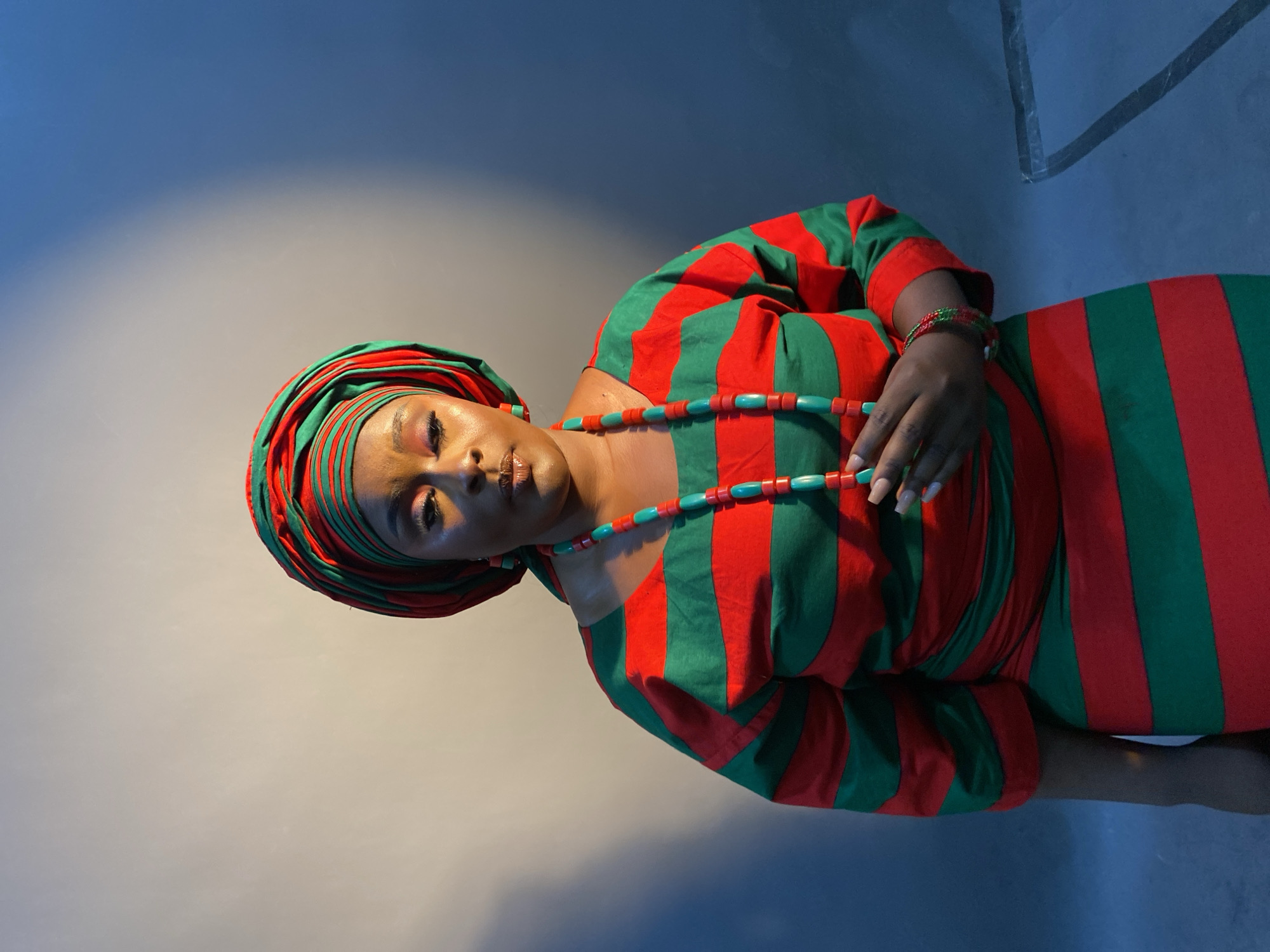 A woman is seen lying on her right side, adorned in a vibrant red and green striped dress and matching headwrap. She wears a necklace with blue beads and a matching bracelet. The scene takes place indoors, set against a gradient backdrop shifting from light gray to deep blue. Specific location in Gura Top, Nigeria is not discernable from the image.