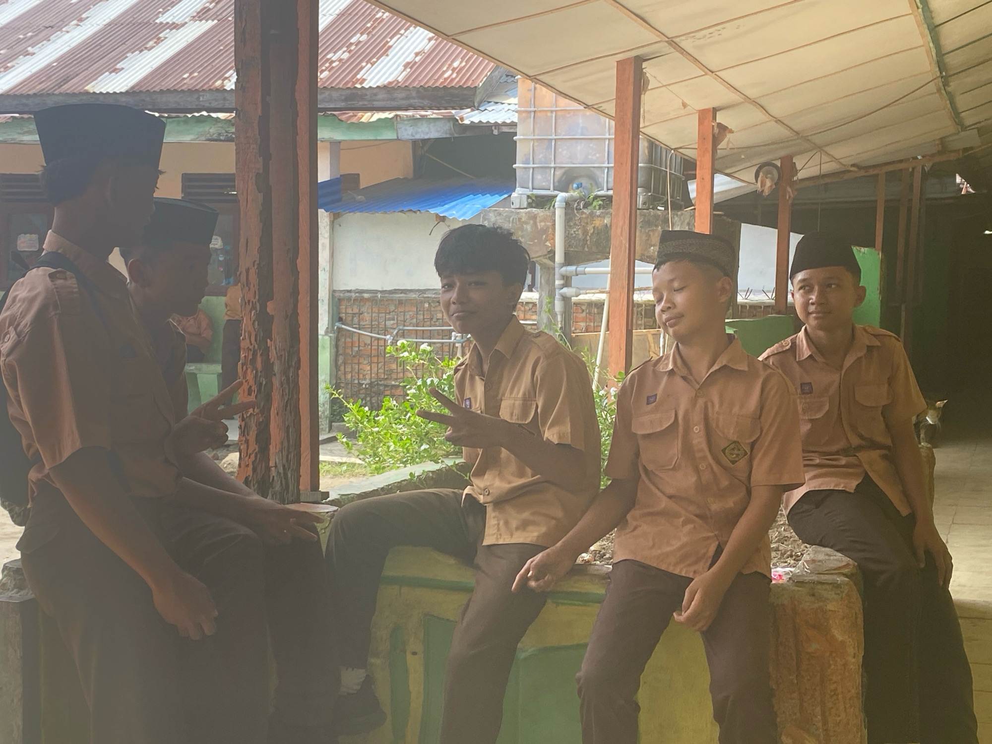 In Karang Gading, Indonesia, five young boys in uniform are gathered on a raised concrete structure, possibly a ledge or low wall, outdoors. Their uniforms consist of light brown short-sleeved shirts with pockets and dark brown pants. Most wear traditional skullcaps, one black and two patterned. The boy on the far left, partially obscured, wears a black skullcap. Next to him, a boy in a brown shirt and black skullcap looks away. The central figure, a boy with dark hair, is looking towards the camera with a slight smile and wearing a black skullcap. To his right, a boy in a brown shirt and patterned skullcap is also looking forward, his eyes partially closed. The boy on the far right, wearing a black skullcap, is looking to the left of the camera. One boy in the center has his hand raised, making a peace sign.
The environment suggests a school or community setting. In the background, a corrugated metal roof is visible, supported by wooden beams. A large plastic water tank is also present, along with some greenery and brickwork. The scene appears to be under a covered walkway or awning, with a light-colored ceiling. The lighting is natural and somewhat diffused, indicating it's daytime. The overall atmosphere is casual and relaxed.