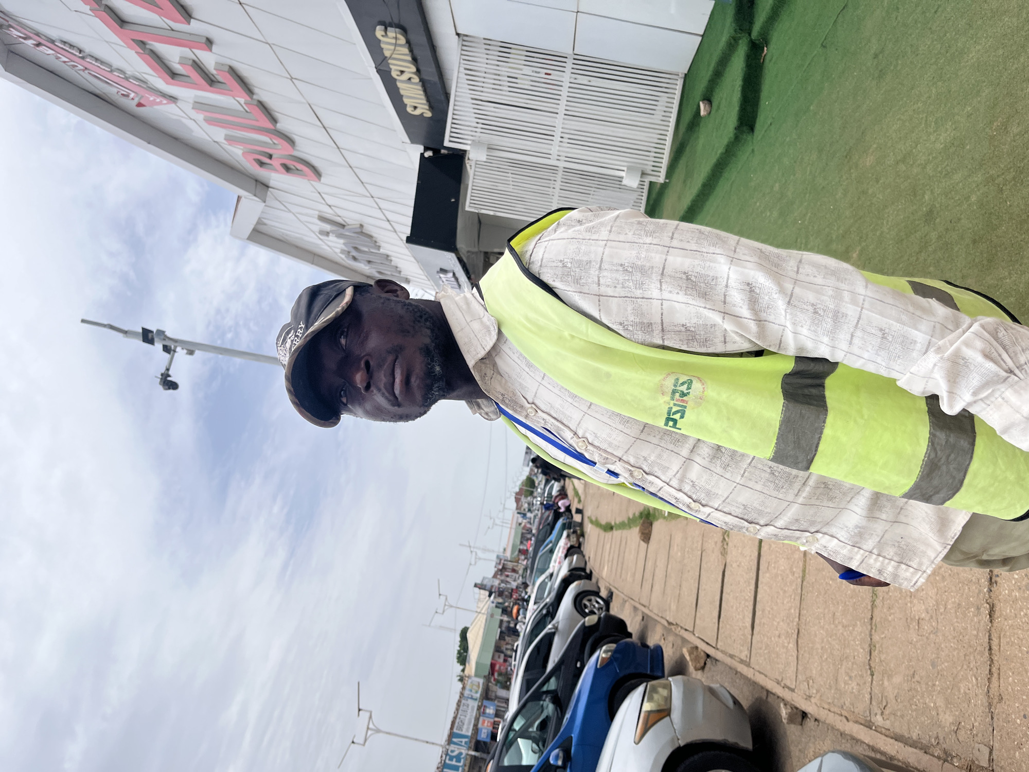 In Jos, Nigeria, a Black man wearing a cap and a high-visibility vest stands in front of a building with a sign that reads "BULERTA." The man is looking upwards and to the right. Behind him, a row of parked cars lines a sidewalk, and a street with shops and other buildings stretches into the distance. The sky is overcast.